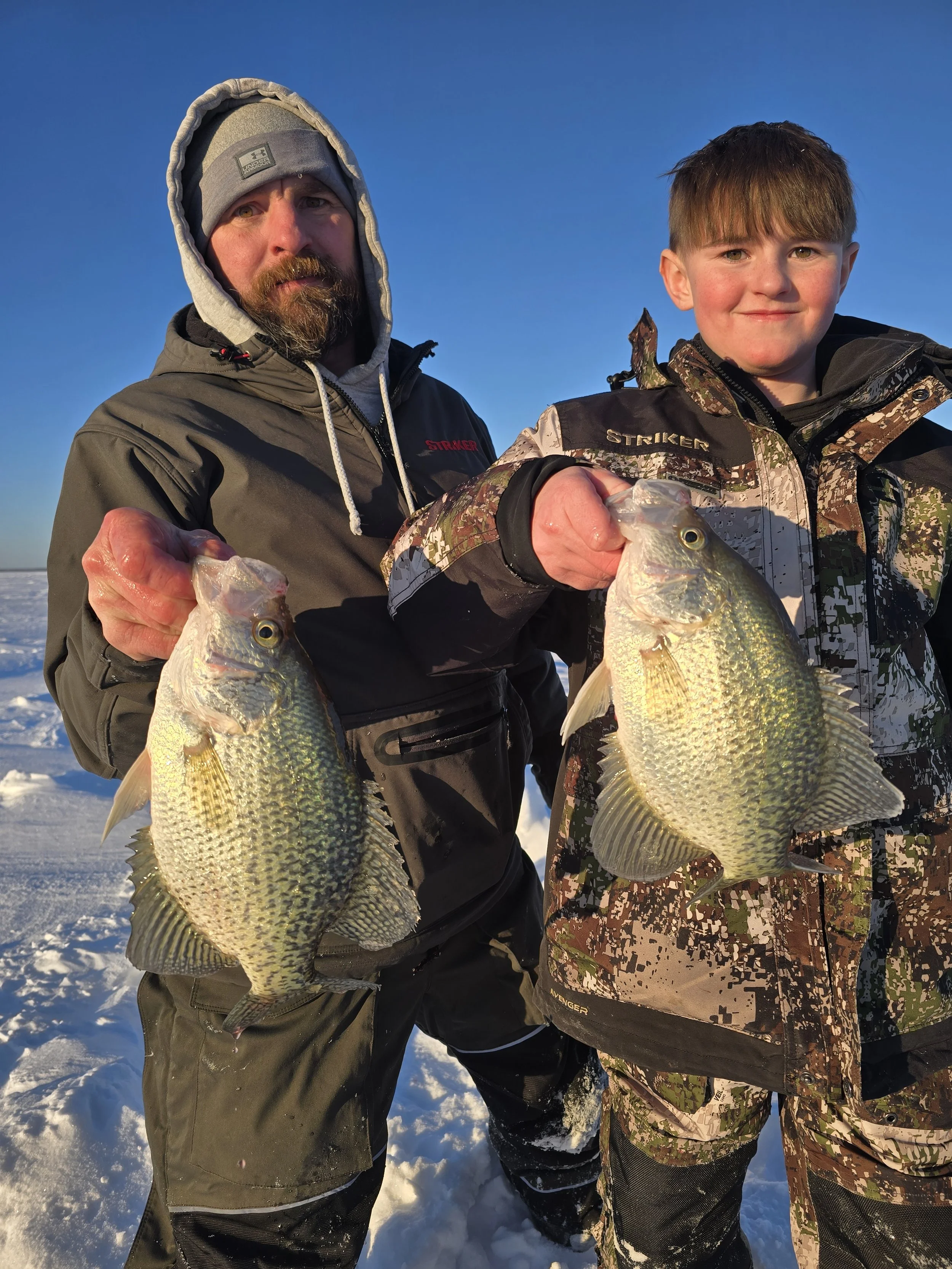 Two men holding large fish on snowy surface, with bright blue sky in background.