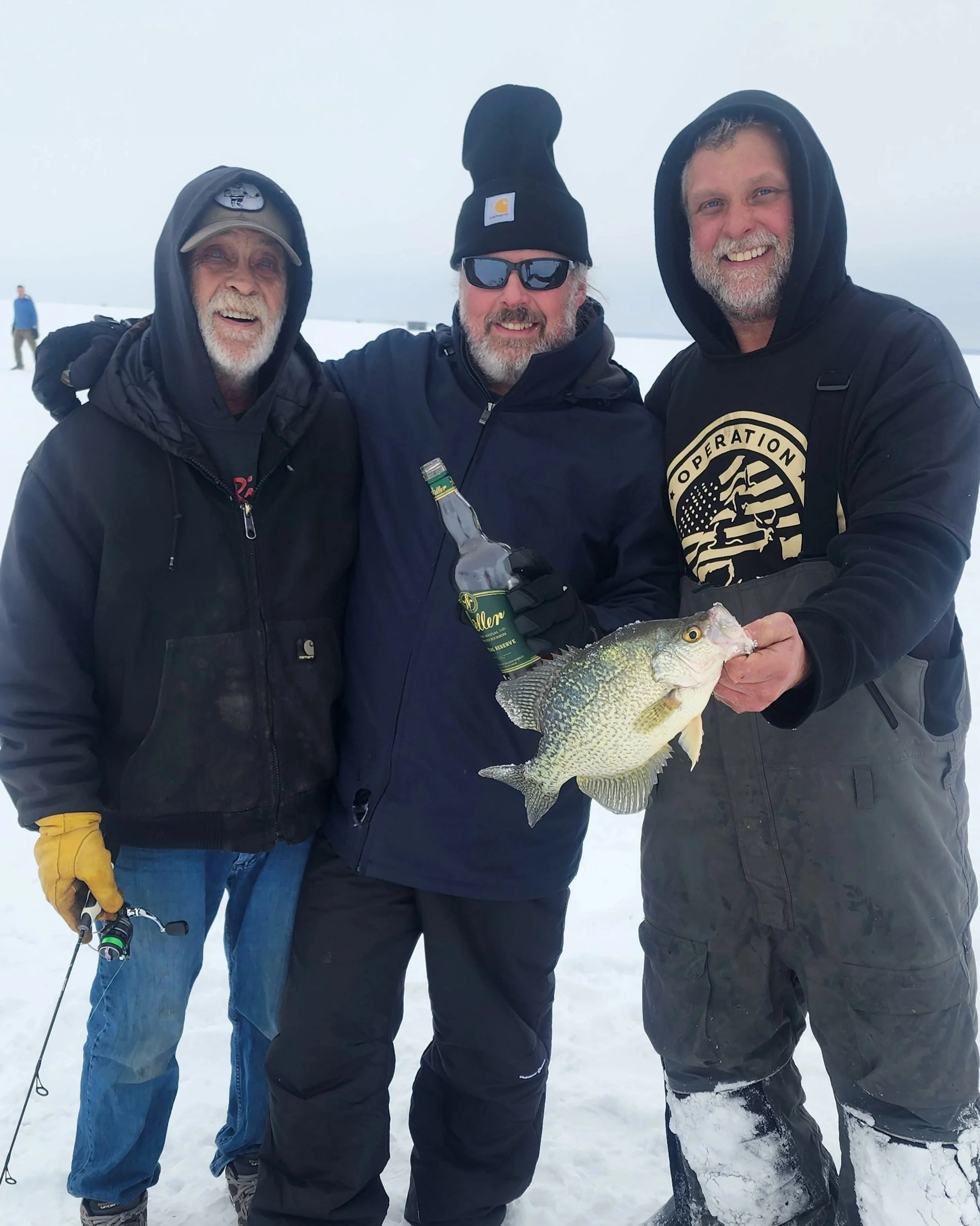 Three men standing outdoors in snowy weather, dressed in winter clothing. The man on the right is holding a large fish. They are smiling and appear to be enjoying a fishing trip.