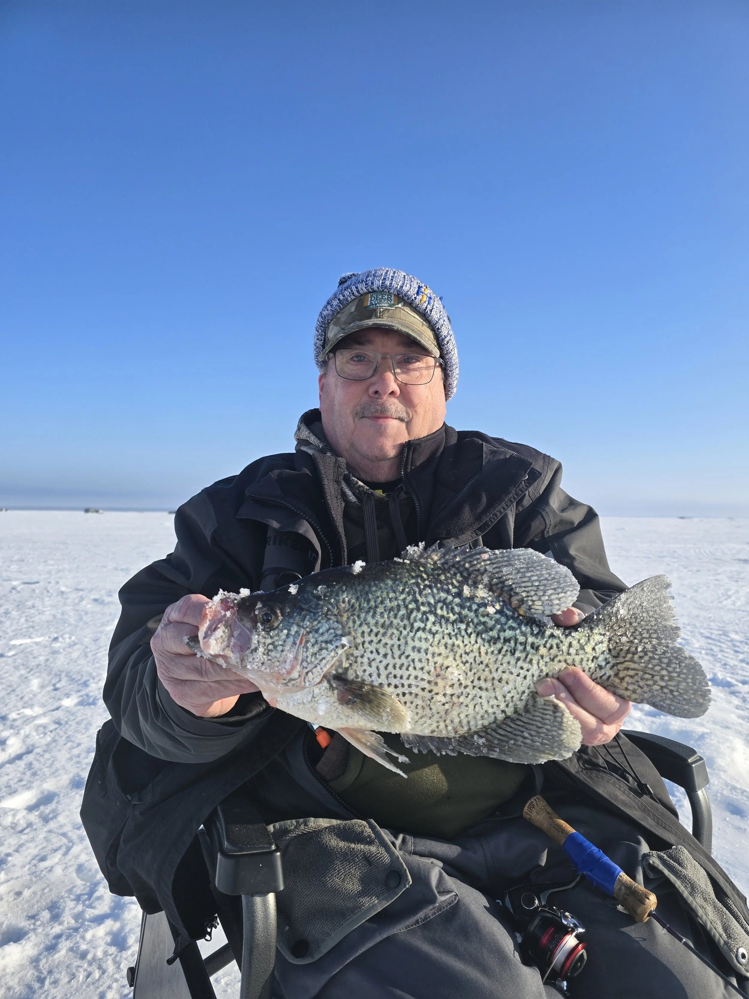 A man in winter clothing and glasses holding a large fish, sitting in a wheelchair on a snow-covered landscape with a clear blue sky.