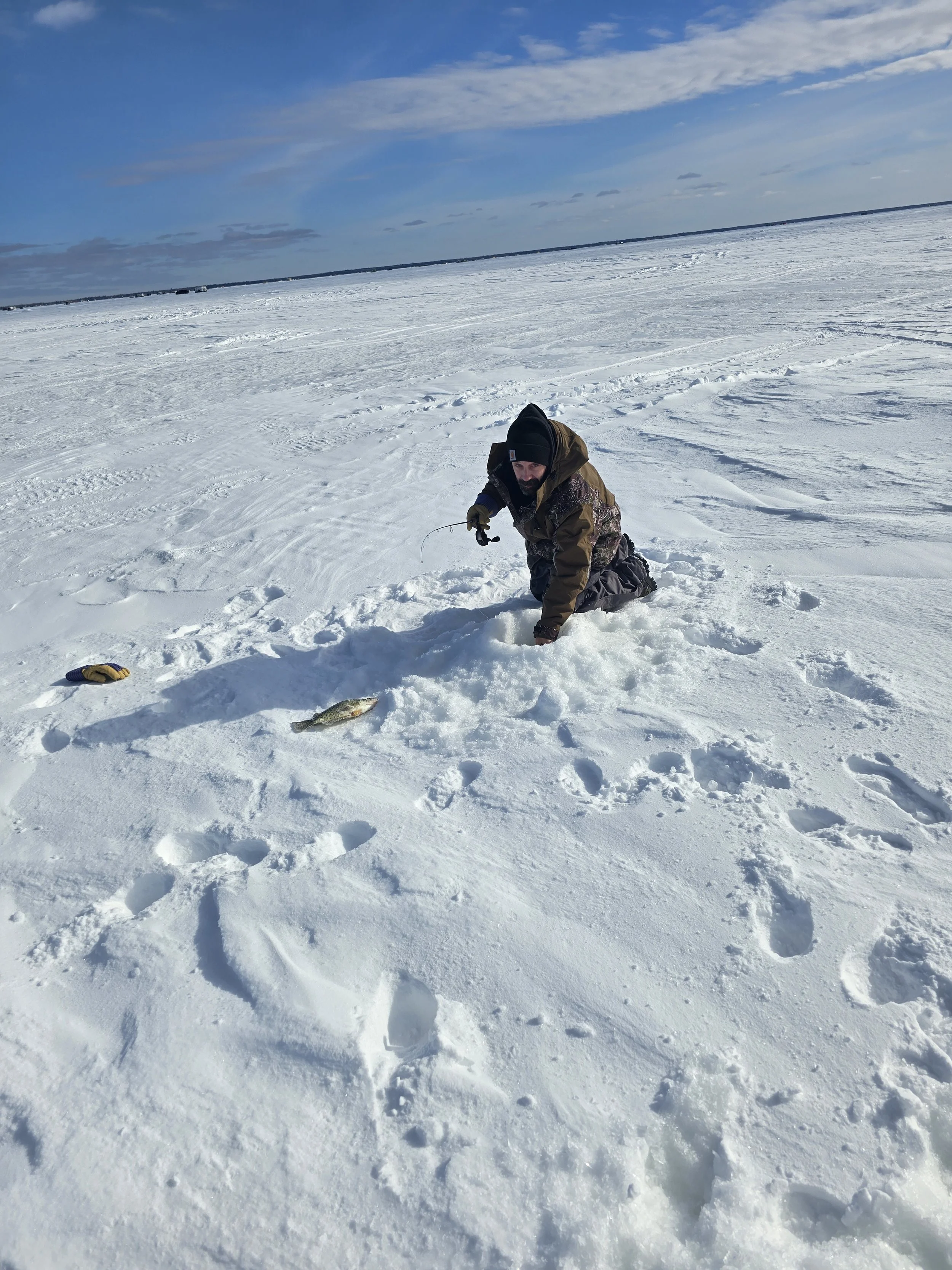 A man kneeling on snow-covered ice holding a fishing rod with a fish in the snow nearby. The background shows a vast frozen landscape under a partly cloudy sky.