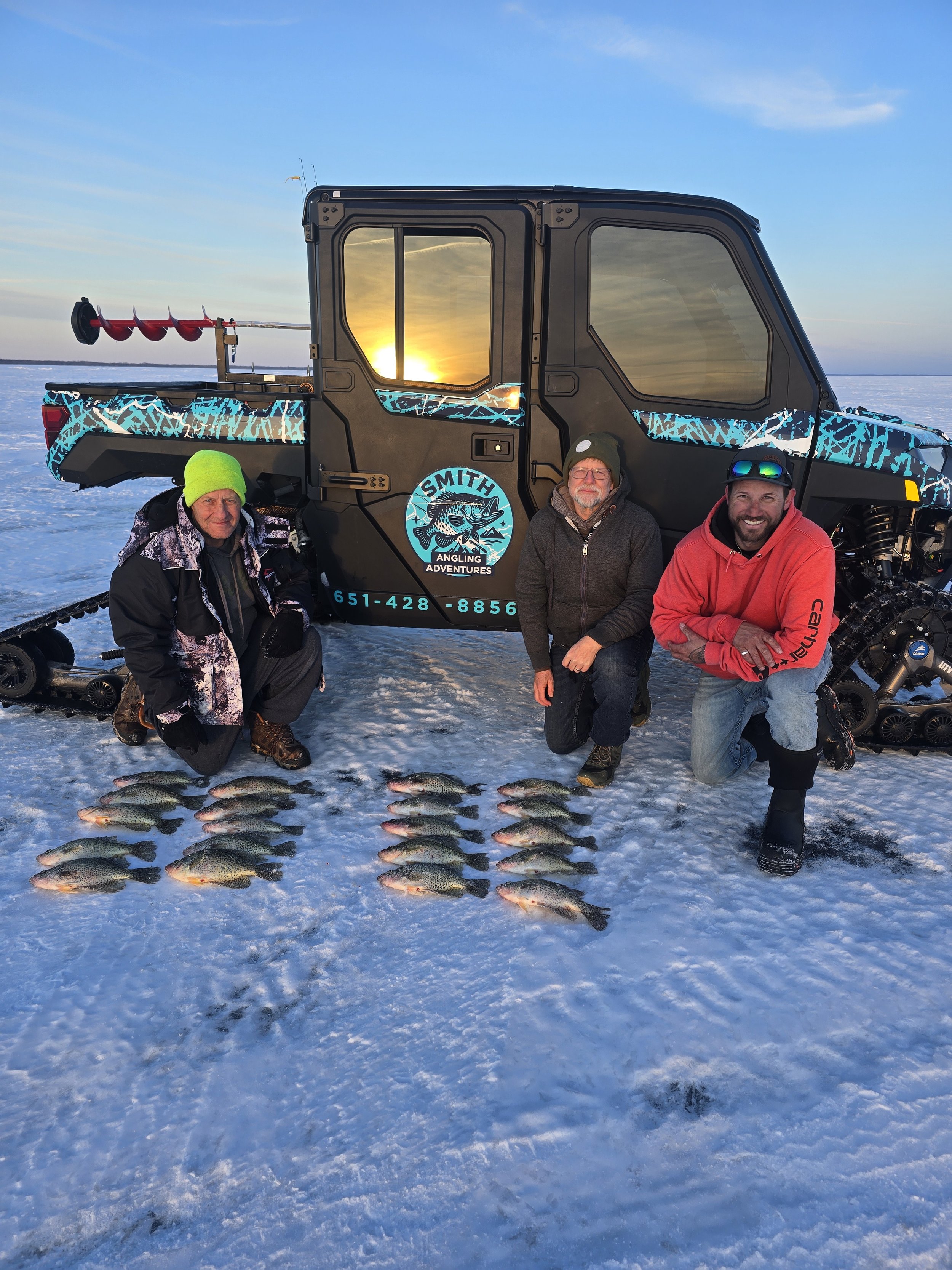 Three men kneeling on snow in front of a snow-covered landscape with a snowmobile and a large vehicle with a logo for Smith Angling Adventures, displaying caught fish, during sunset.