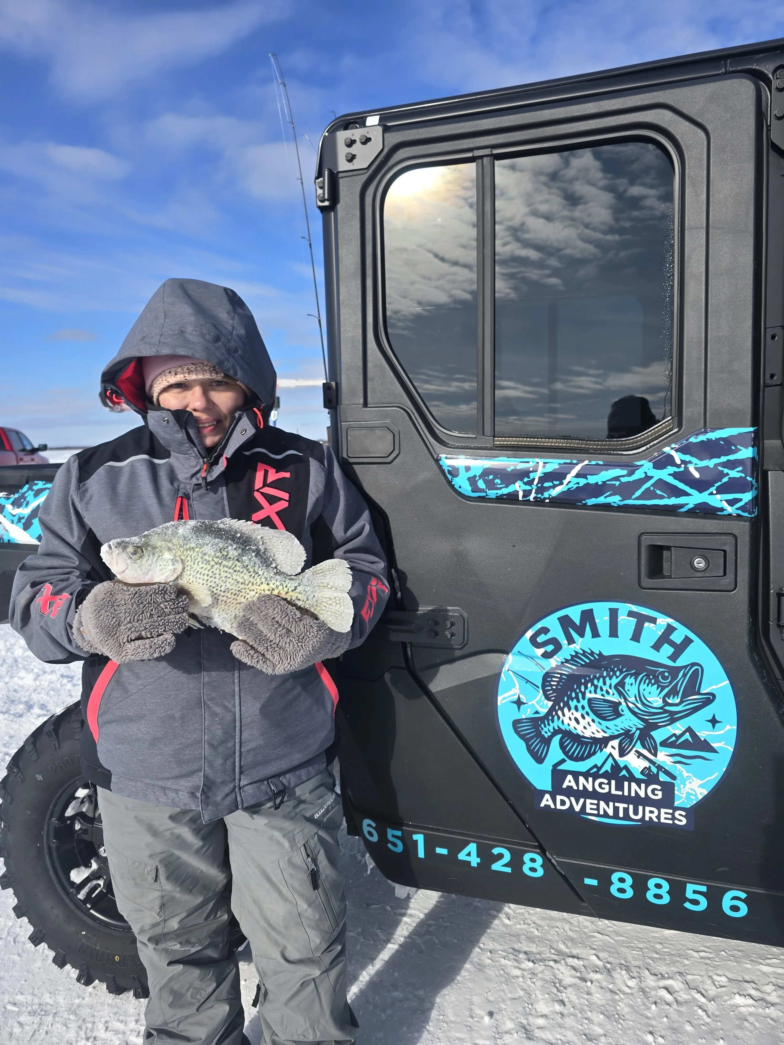 A young boy wearing winter clothing, including a hooded jacket and gloves, holding a fish in front of a black vehicle with a logo that says 'Smith Angling Adventures' and a mountain illustration. The background features a blue sky with some clouds an