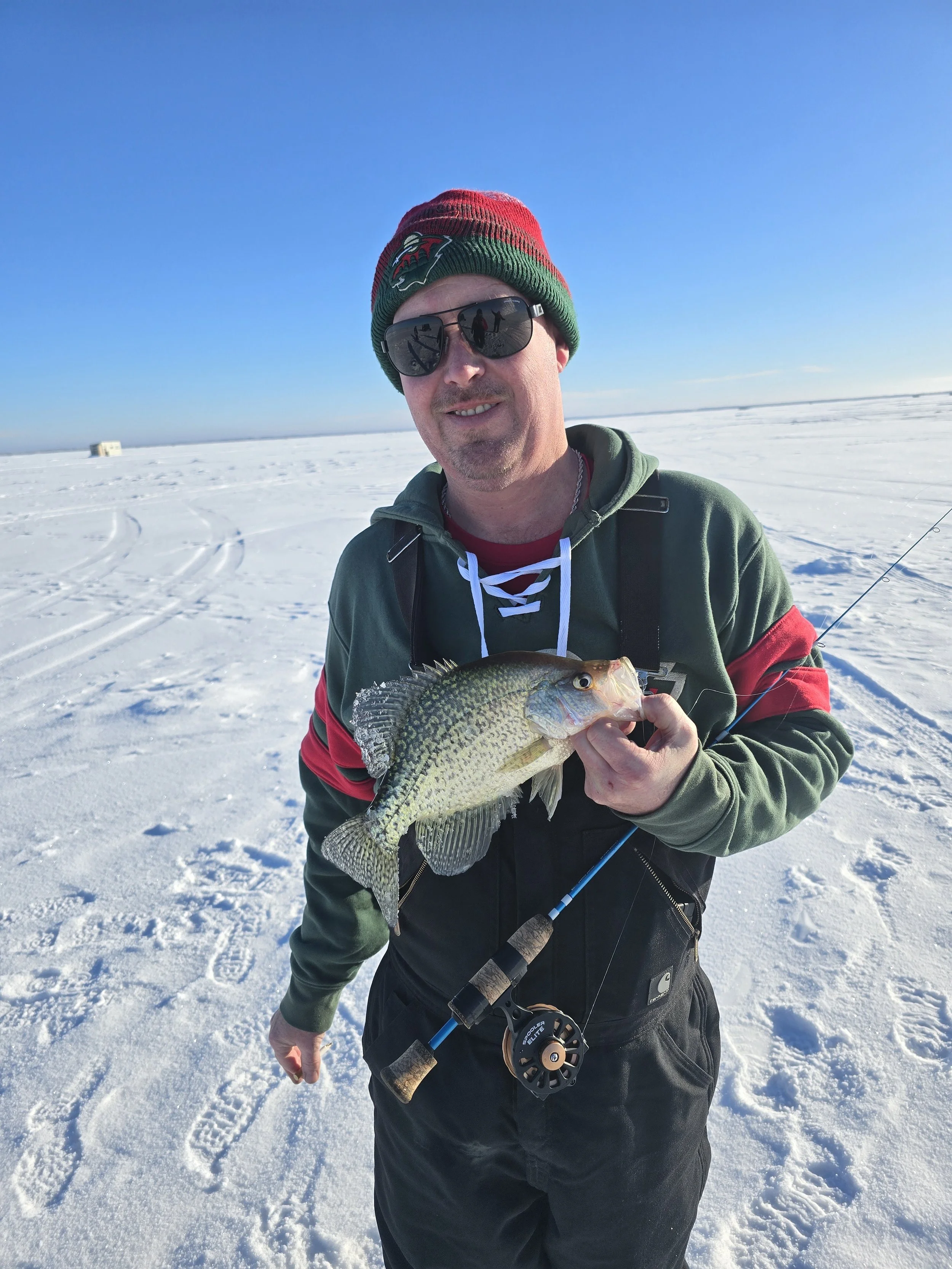 Man in sunglasses and a red and green beanie holding a fish in a snowy landscape with clear blue sky.