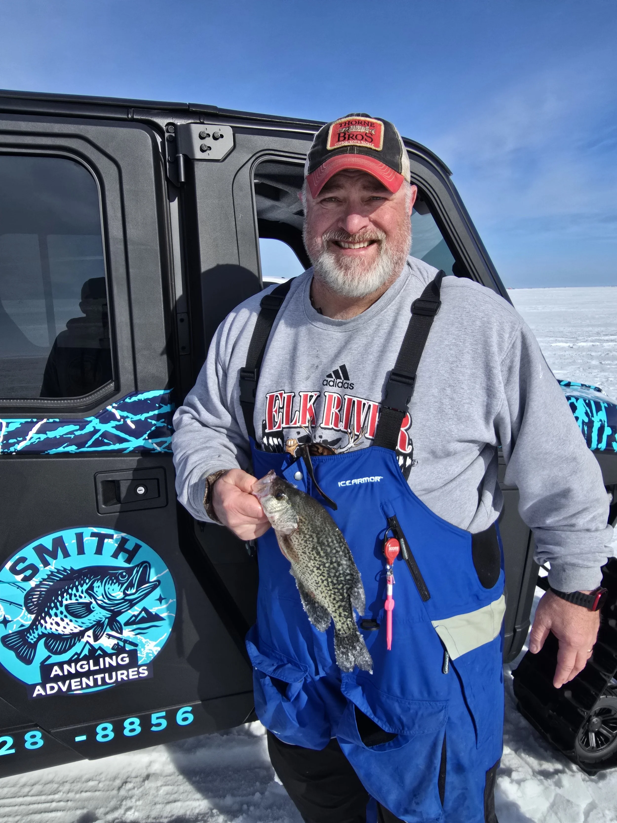 A smiling man in a gray sweatshirt and blue overalls holding a fish, standing in front of a black vehicle with the logo 'Smith Angling Adventures' on it, snow on the ground, and a clear blue sky.