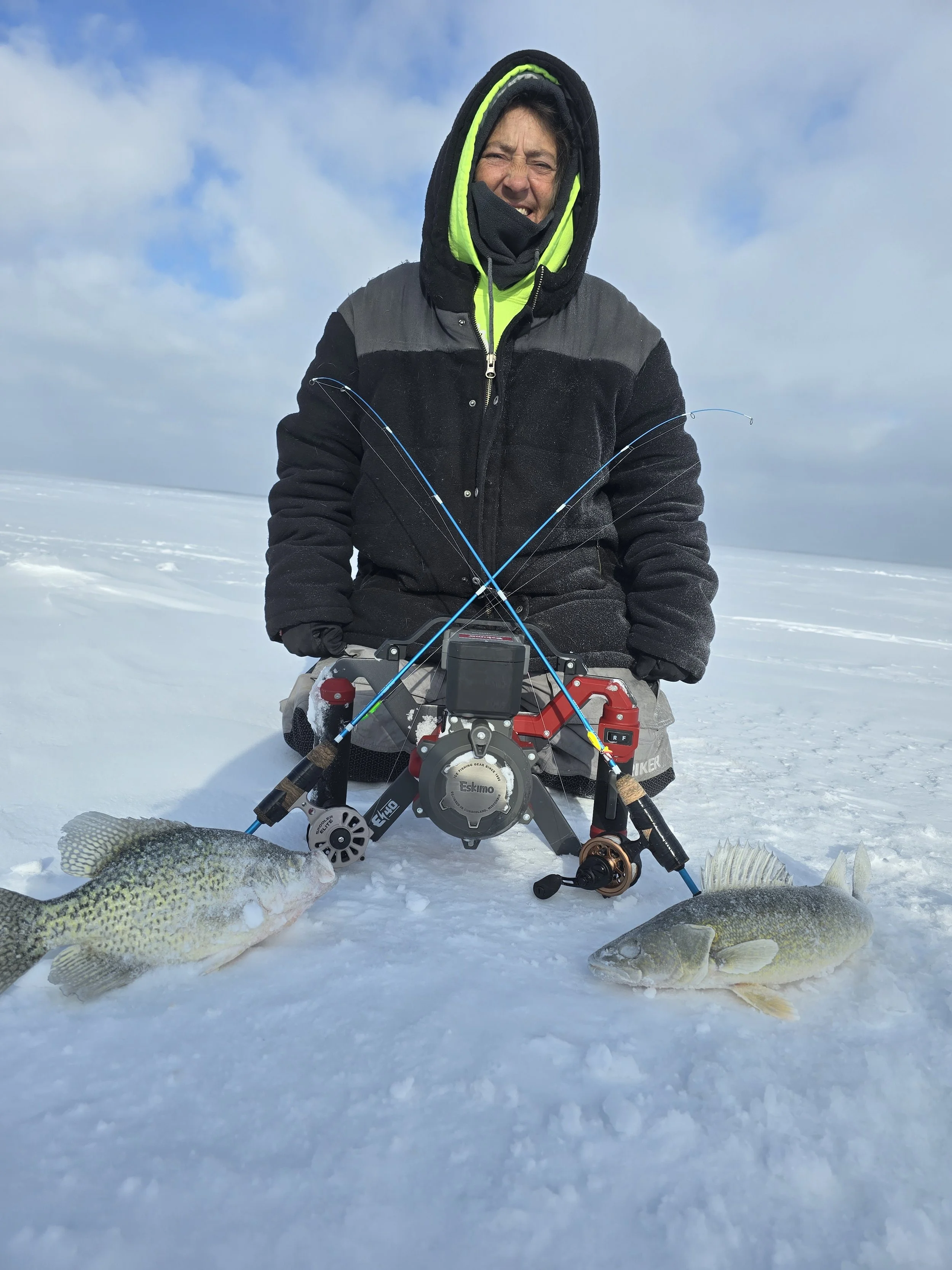 Person in winter clothing sitting on snow with four caught fish laid out in front, holding fishing rods with a snowy landscape and partly cloudy sky in the background.