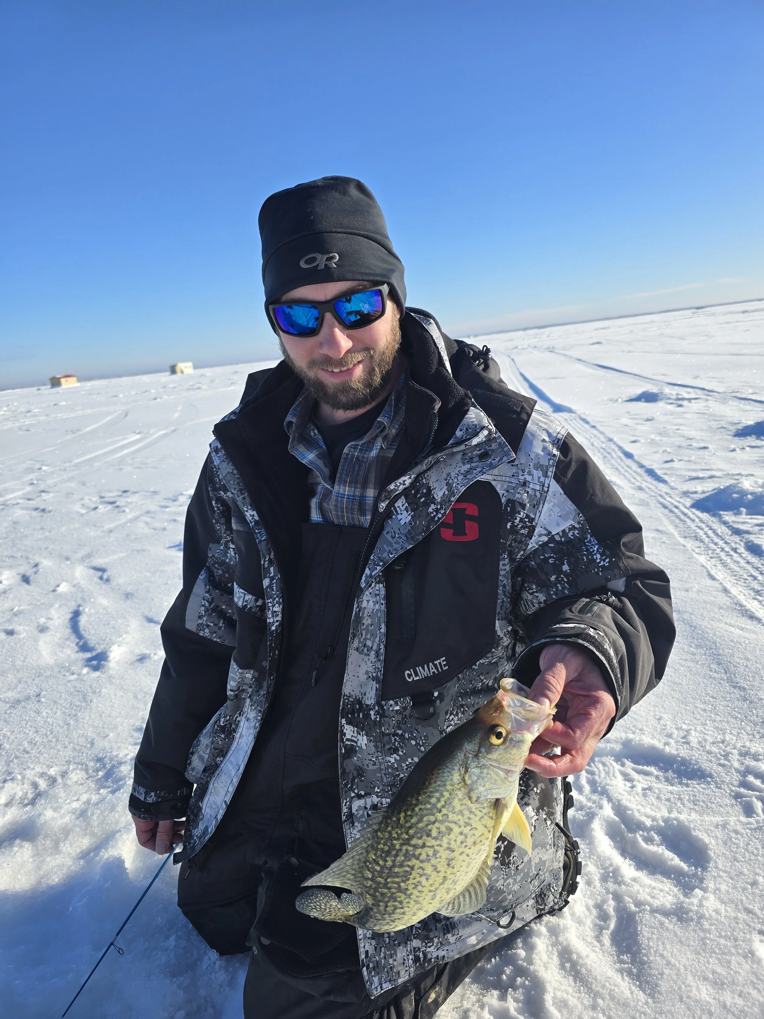 A man dressed in winter gear holding a large fish on a snowy landscape during daytime with a clear blue sky in the background.