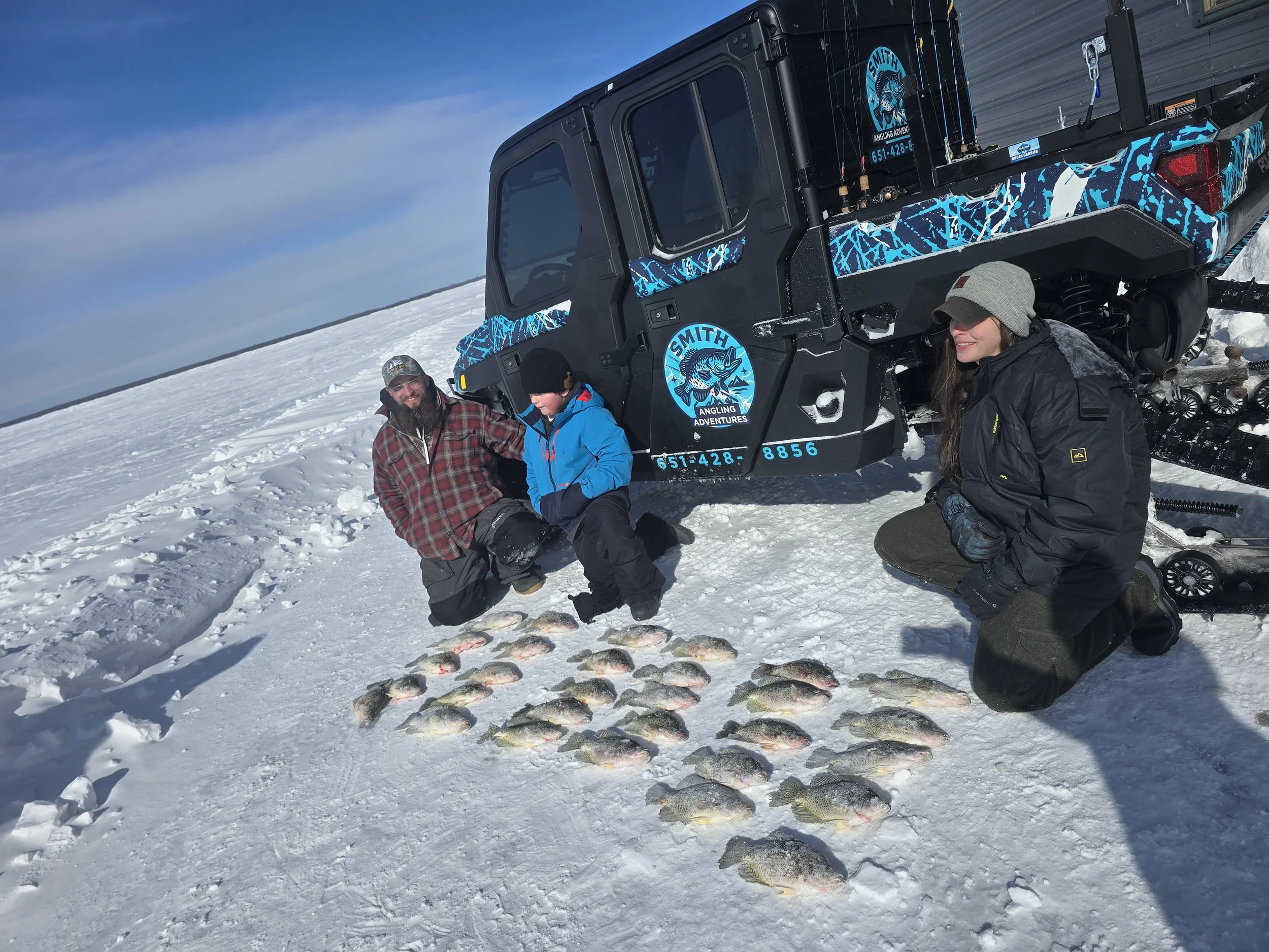 A group of three people on a snowy landscape with a black vehicle in the background, displaying a logo for Smith Angling Adventures. They are posing with several fish laid out on the snow, dressed in winter clothing, and smiling.
