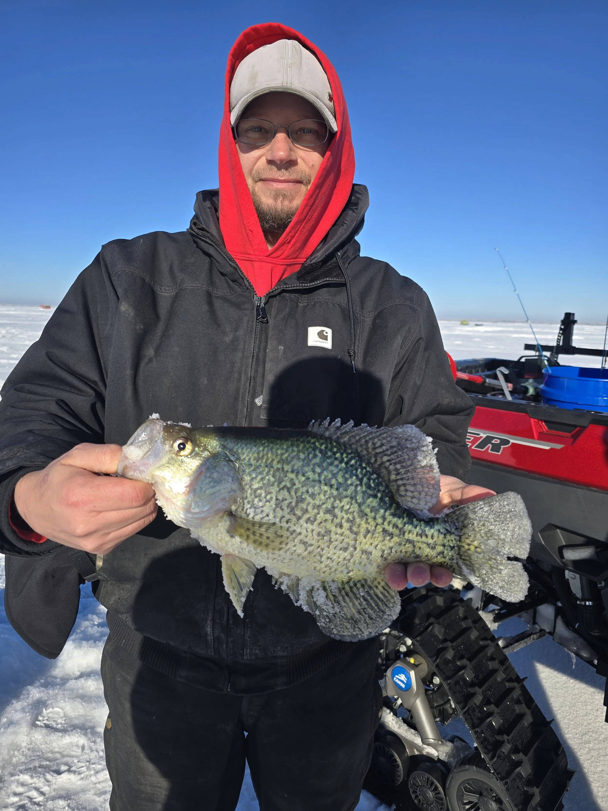 A man holding a large fish on snow-covered ground with a snowmobile and fishing gear in the background.
