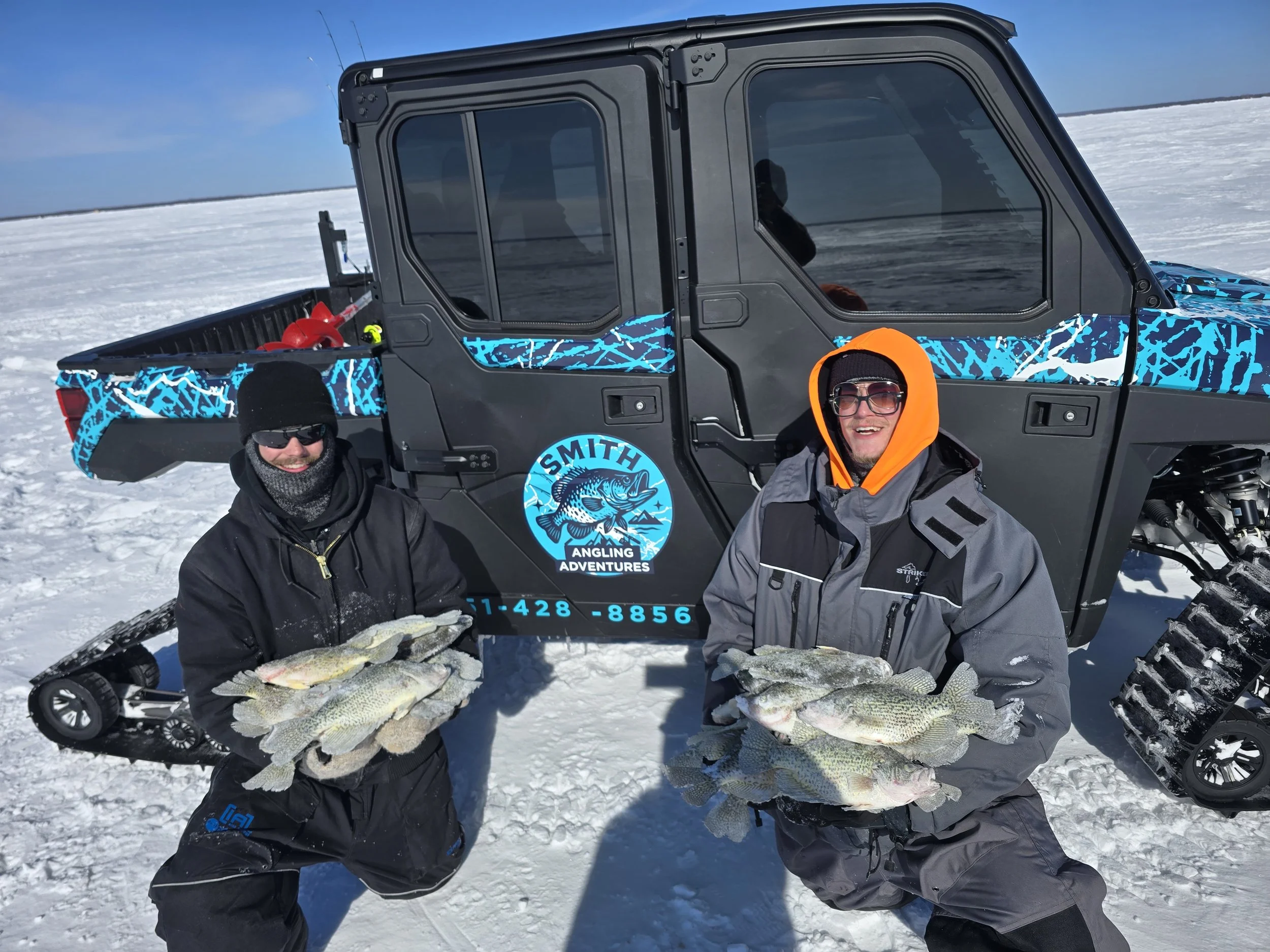 Two men in winter clothing holding multiple large fish in front of a side-by-side ATV on snow, with a snowy landscape and clear blue sky in the background.
