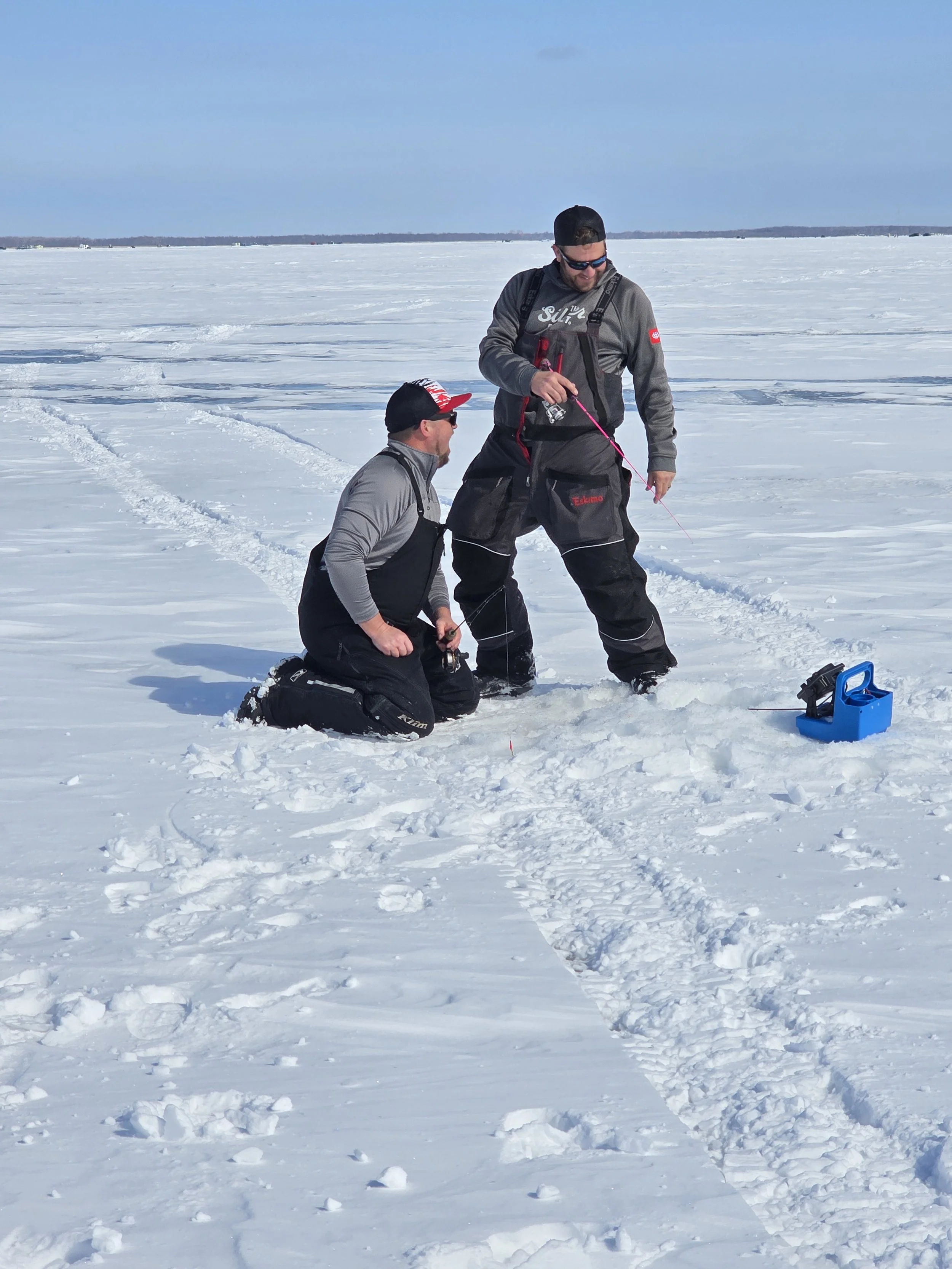 Two men are ice fishing on a large frozen lake with snow-covered ground and tracks in the snow. One man is kneeling and holding a fishing rod, while the other stands holding a fishing pole. There is a blue tackle box on the snow near them. The sky is