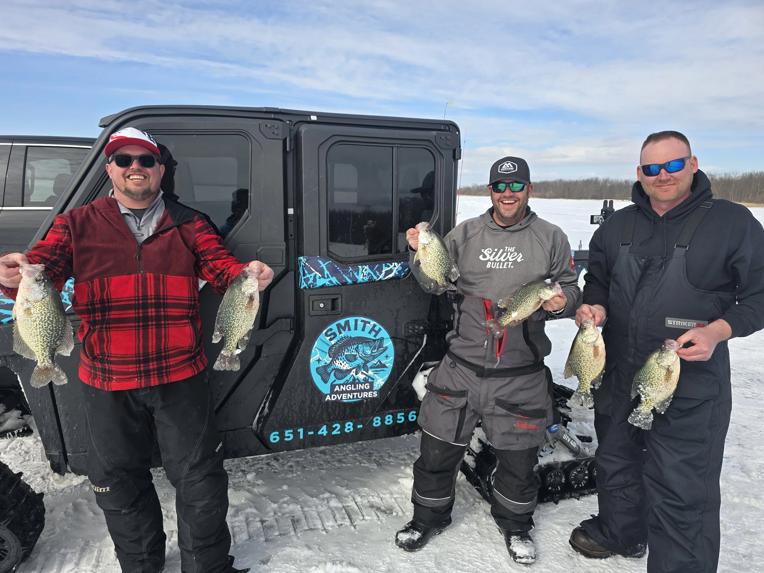 Three men standing in the snow holding fish they caught during a winter fishing trip, with a black off-road vehicle behind them on a snowy landscape.