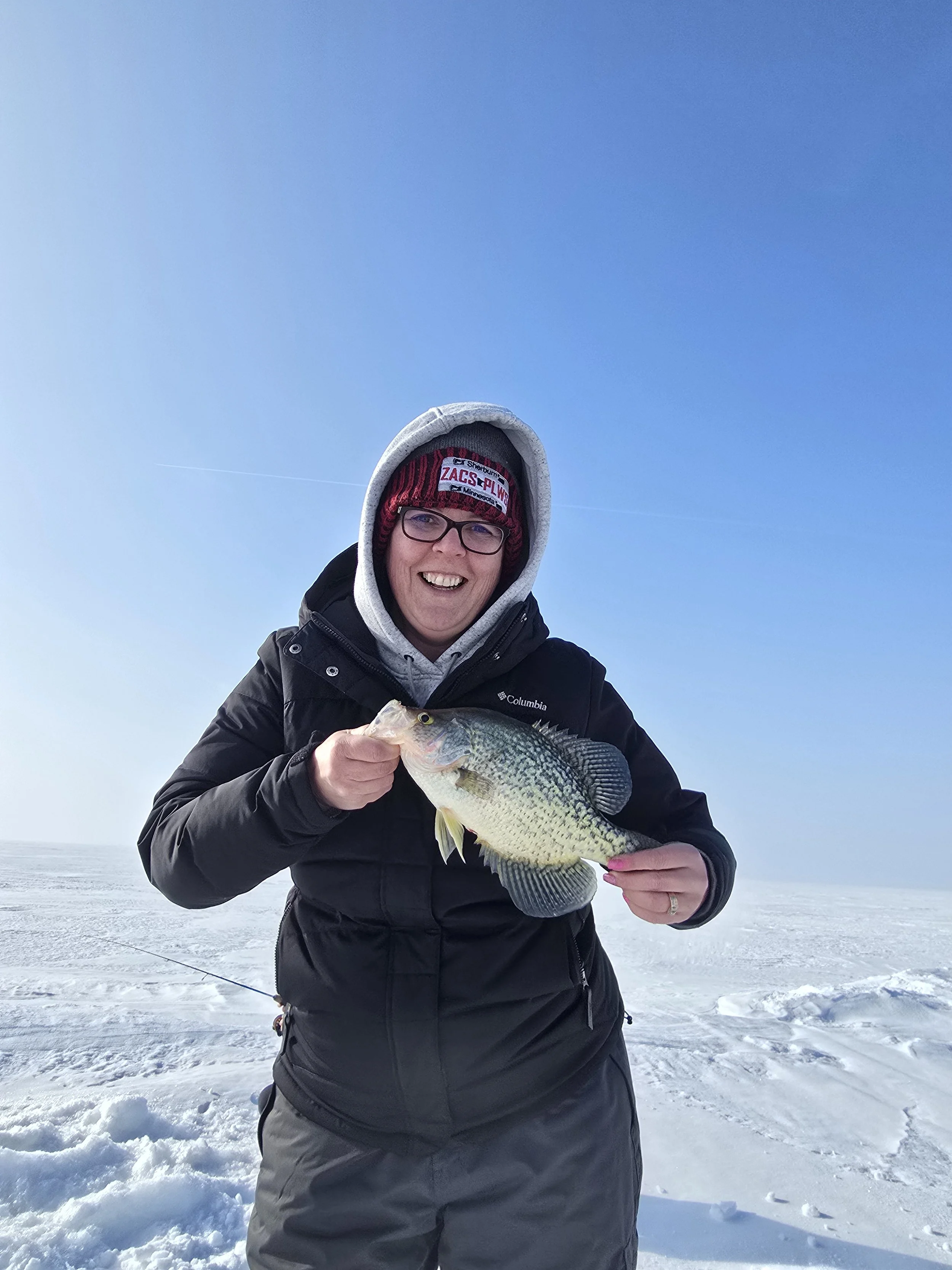 Person in winter clothing holding a fish on a snow-covered surface with clear blue sky in the background.