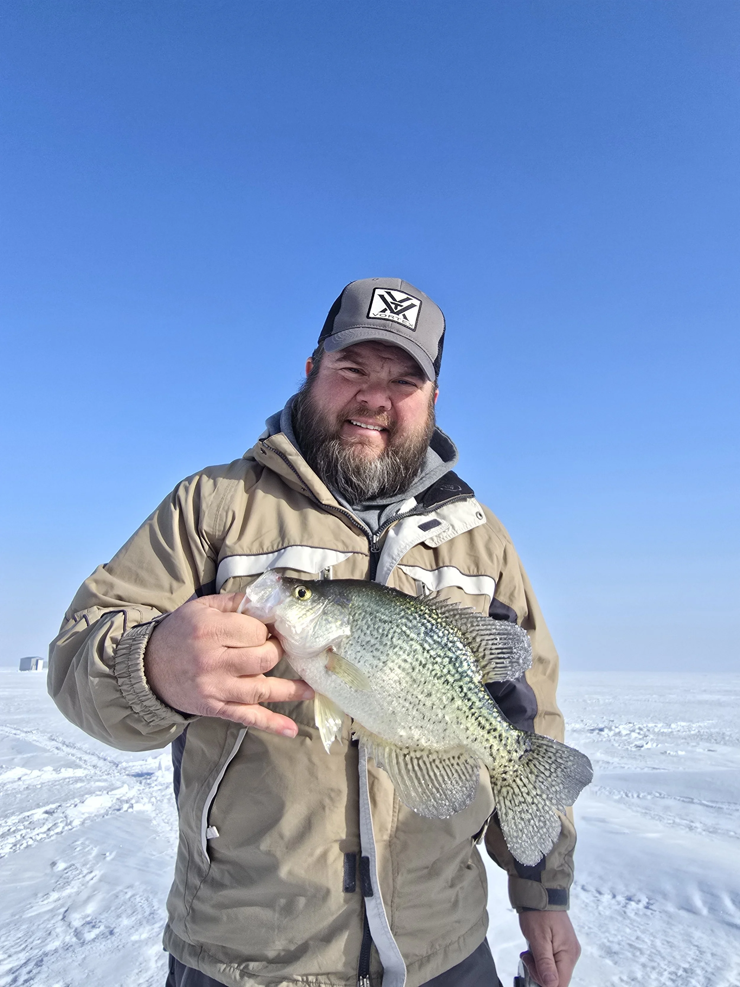 A man standing on ice in winter weather, holding a large fish with greenish and grayish scales. He wears a beige winter jacket and a gray cap with a black and white logo. The background shows a clear blue sky and snow-covered ground.