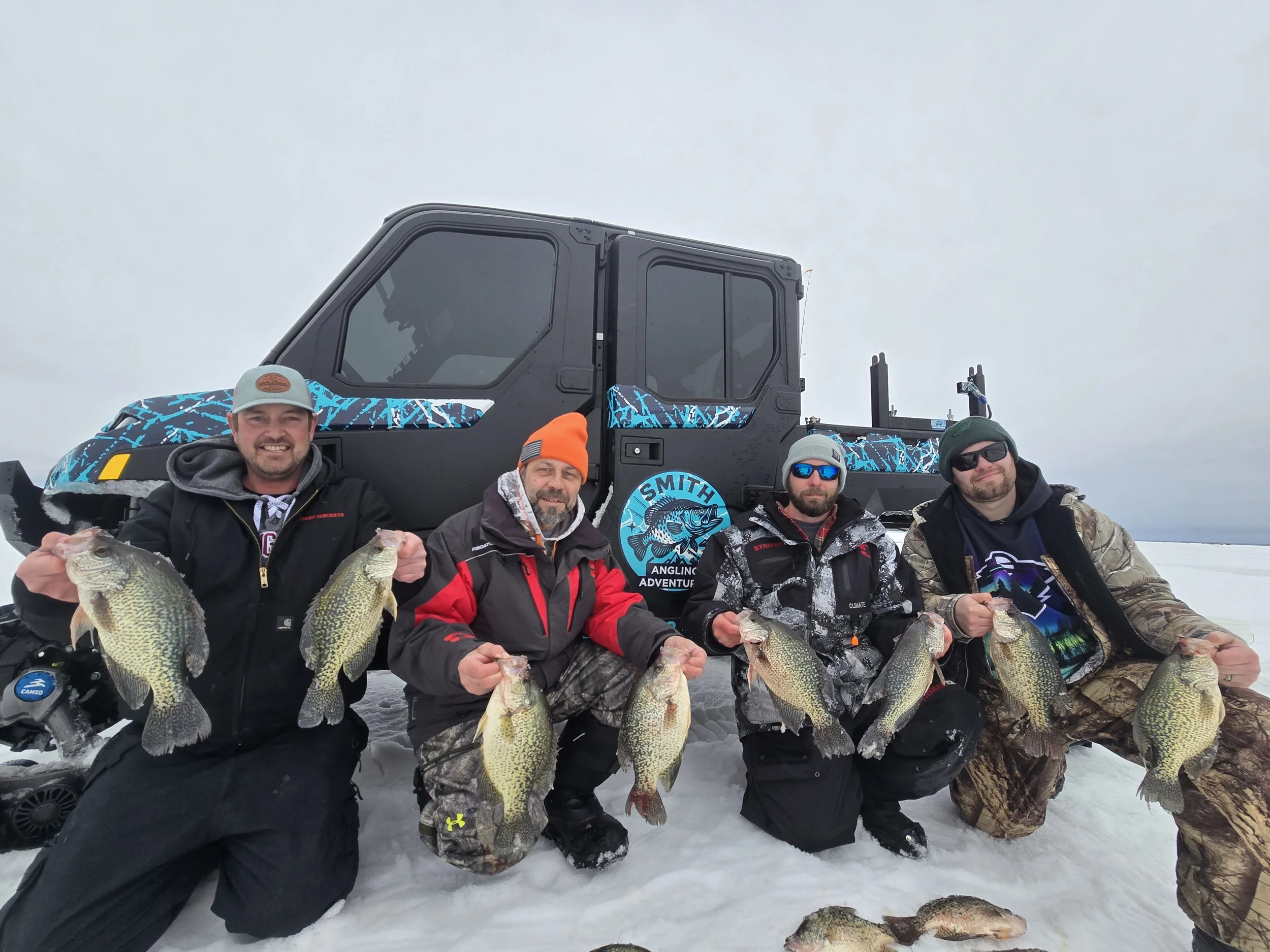 Group of five men outdoors in winter holding large fish, posing in front of a snow-covered landscape with a black snowmobile behind them. They are dressed in winter clothing including jackets, hats, and sunglasses.