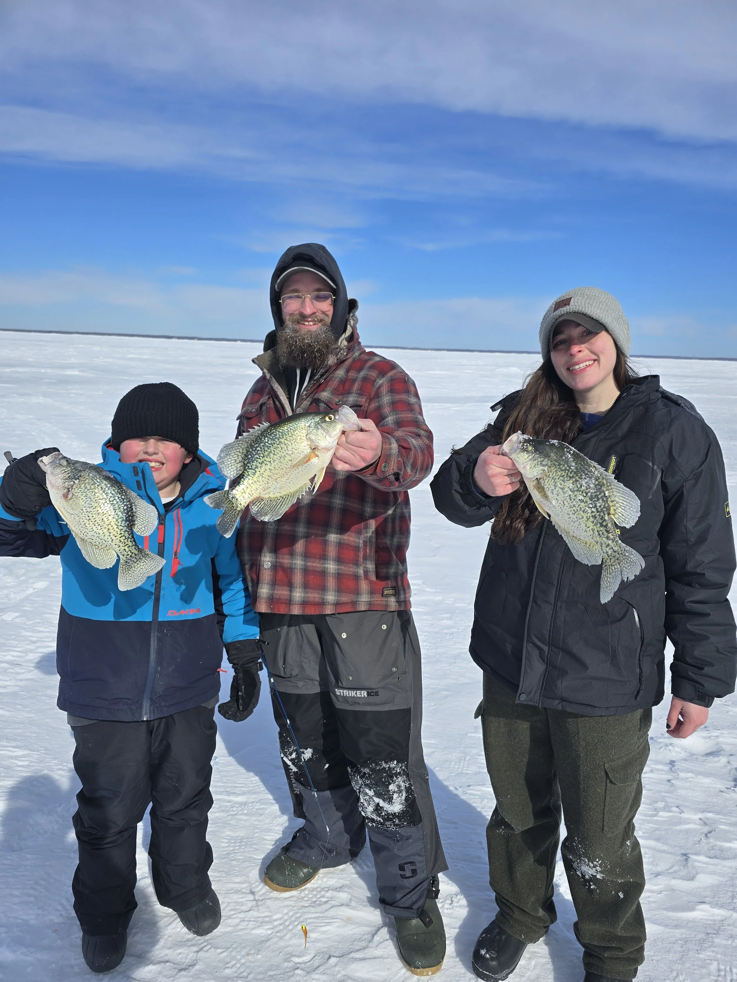 Three people, two adults and one child, standing on snow-covered ice in a frozen lake or river, holding large fish they caught. They are dressed warmly in winter clothing, and the sky is partly cloudy.