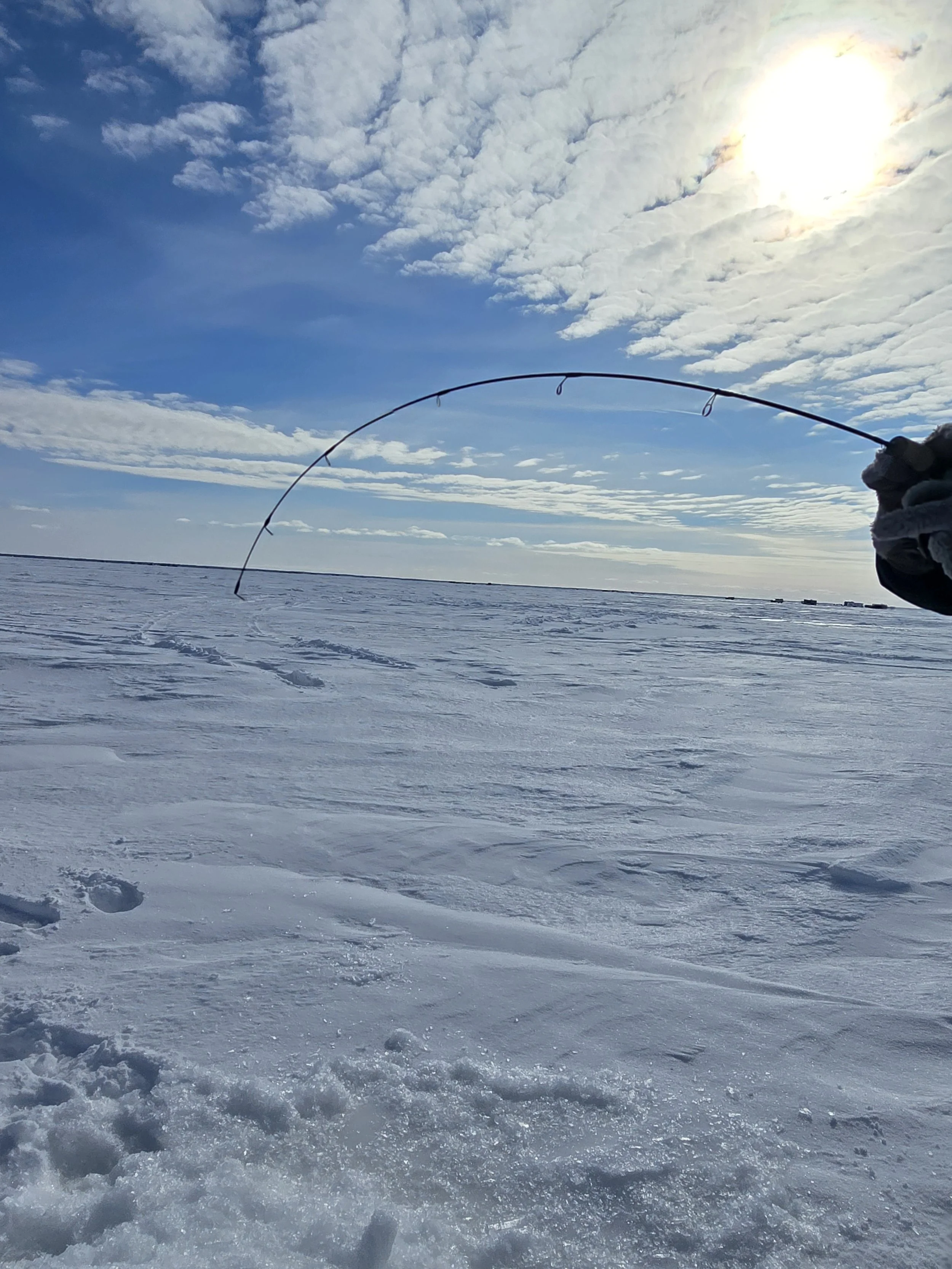 Person fishing on a frozen lake with snow, clouds, and the sun in the sky.