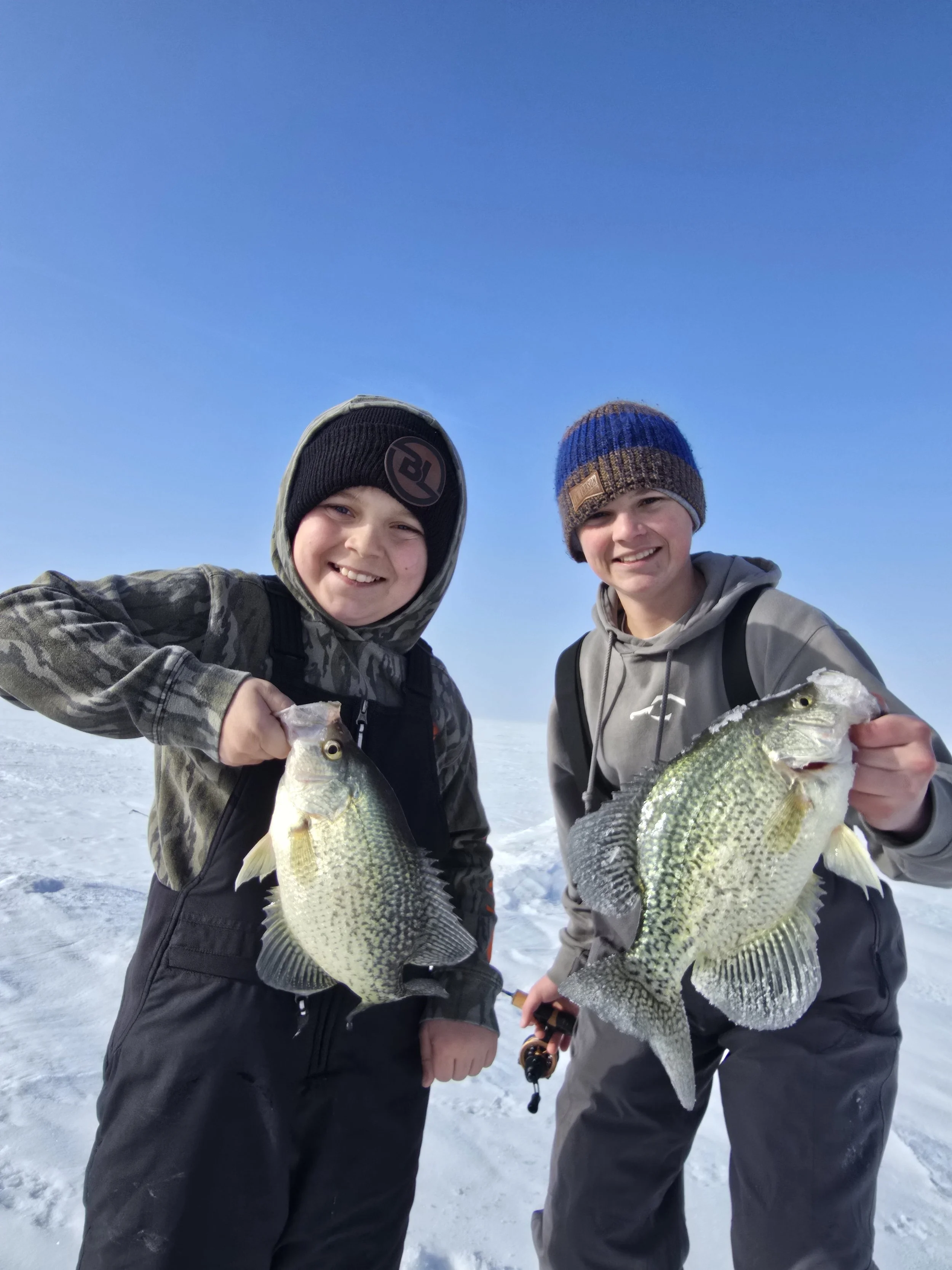 Two young boys in winter clothing on snow-covered ice, each holding a large fish they caught, under a clear blue sky.