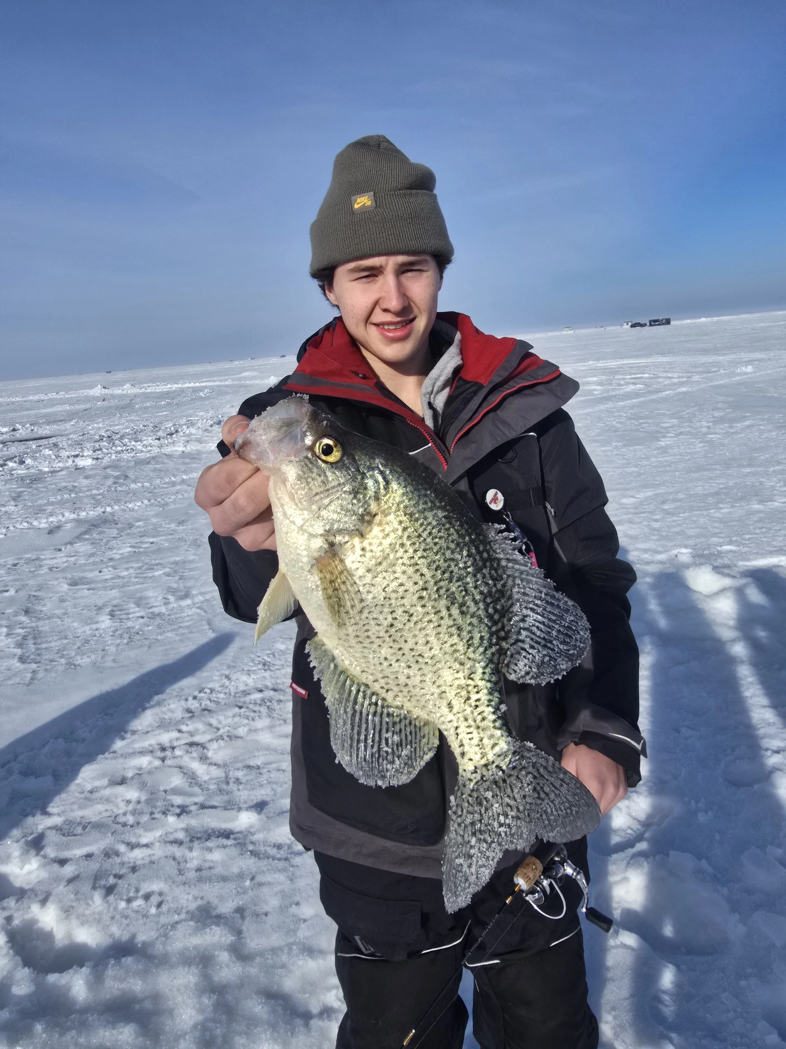 A young man in winter clothing holds a large fish on a snowy, frozen landscape with a clear blue sky and truck tracks in the distance.
