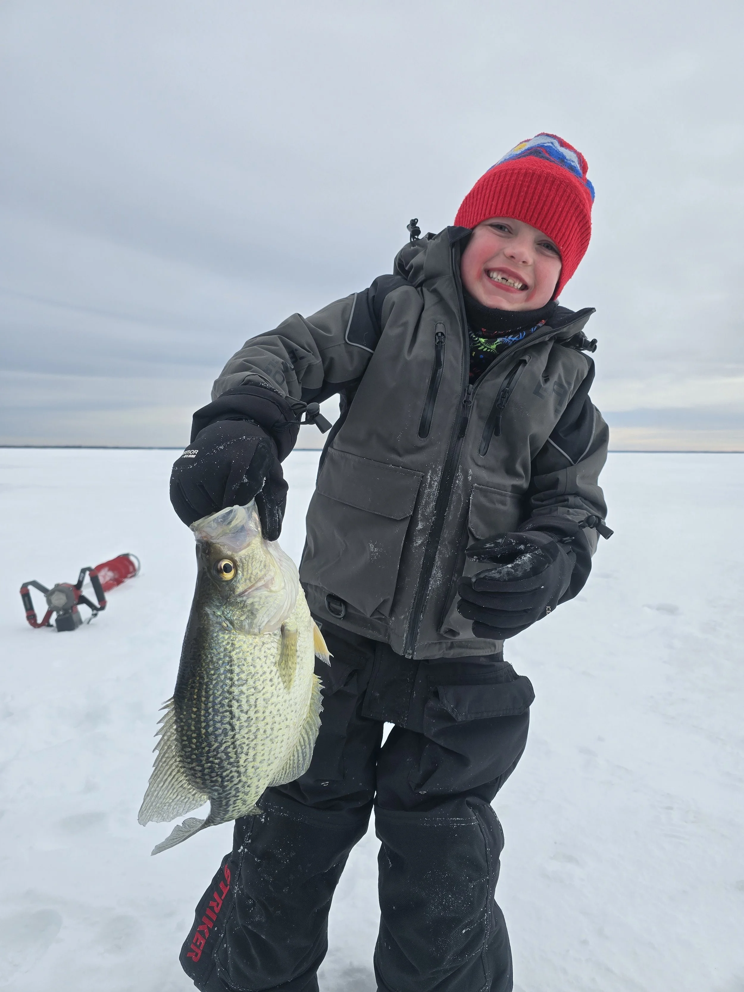 A smiling child in winter clothing holding a large fish on a snowy landscape during daytime.