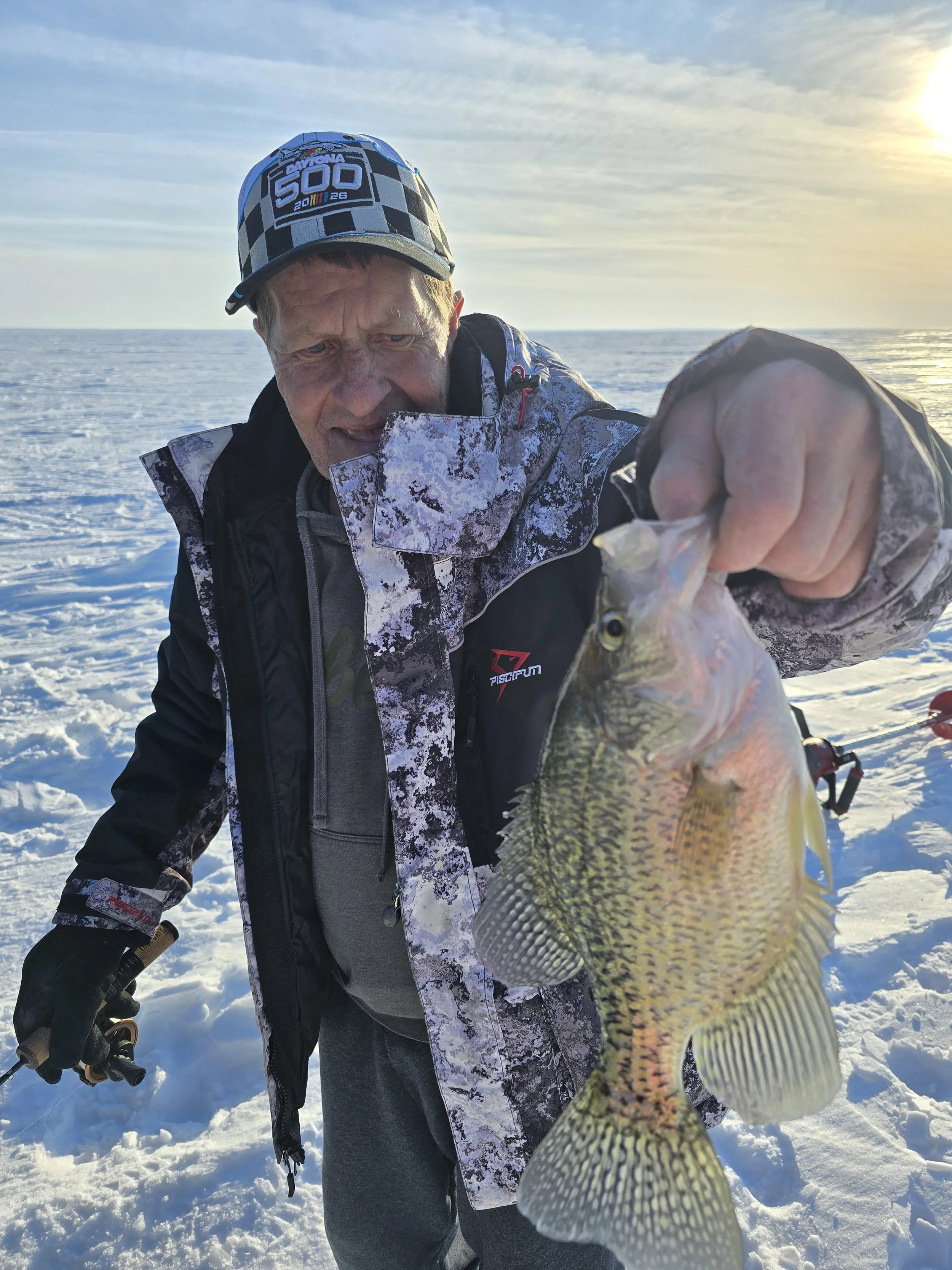 Man holding a fish while ice fishing on a snowy lake during sunset.