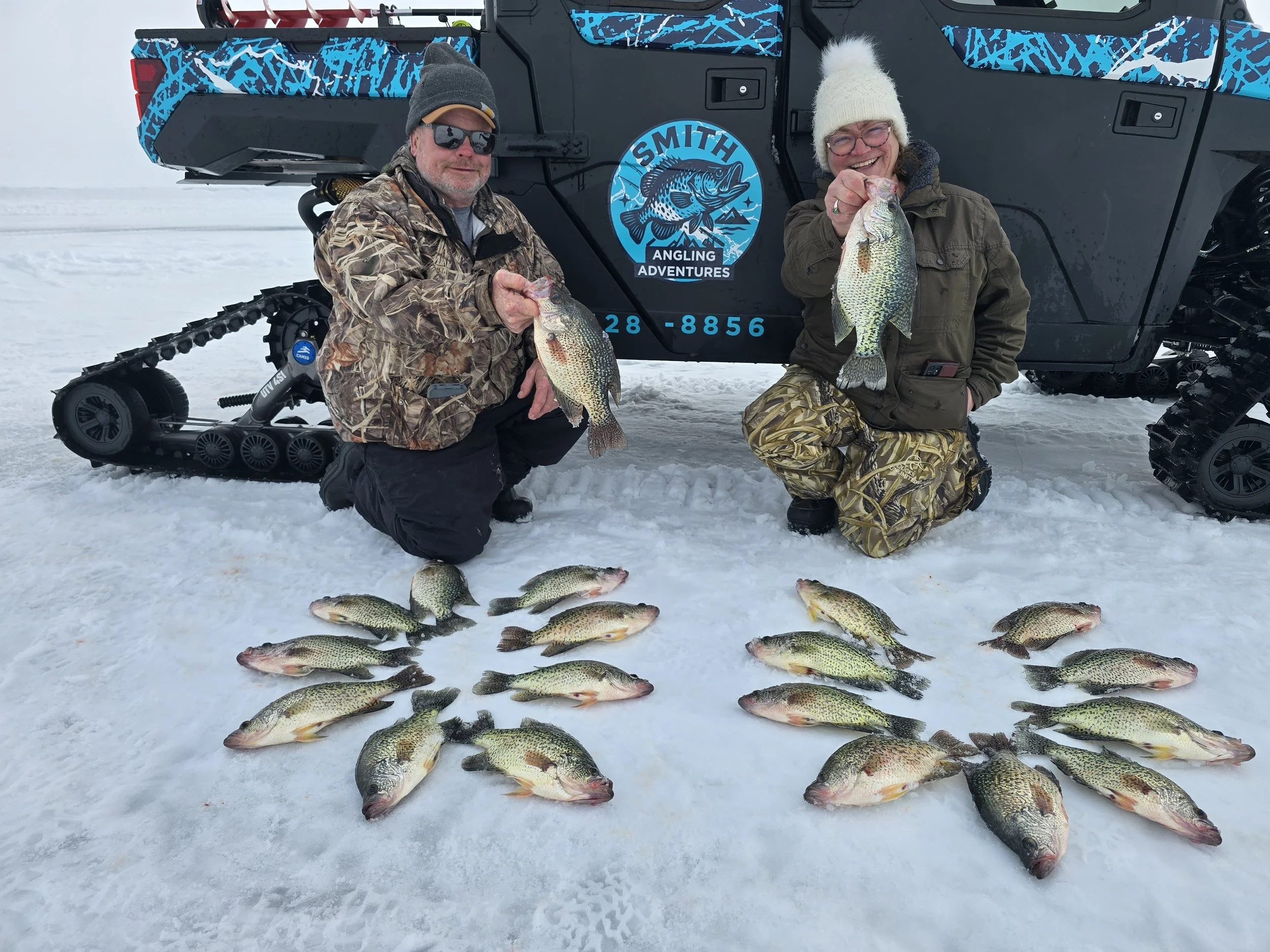 Two people kneeling on snow-covered ground holding fish, with more fish laid out in front, in front of a black vehicle with a blue and black logo that reads 'Smith Angling Adventures.' The person on the left is wearing camouflage clothing and sunglas