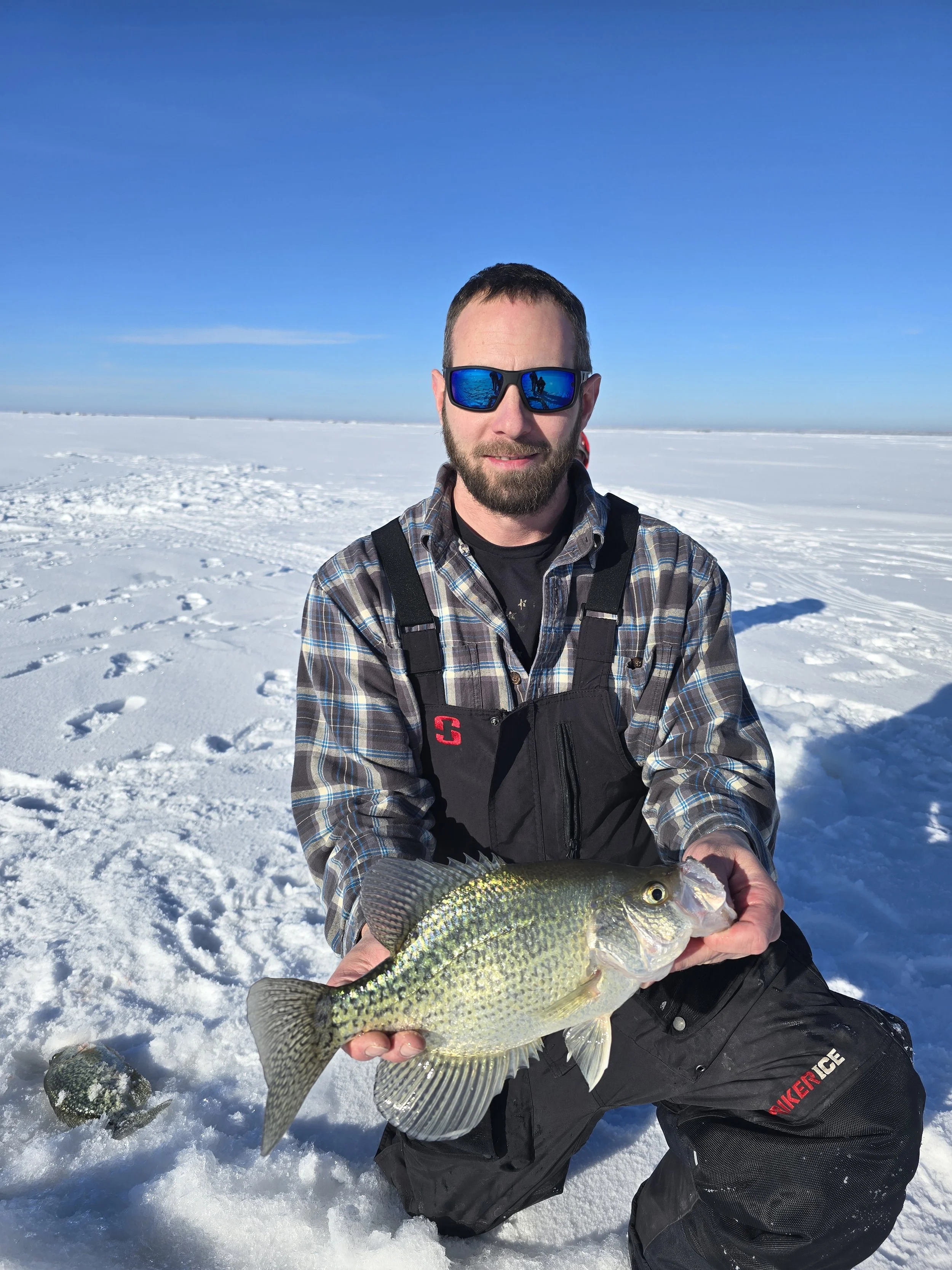 A man wearing a plaid shirt, sunglasses, and a black vest kneels on snow, holding a large fish he caught, with snowy landscape and a clear blue sky in the background.
