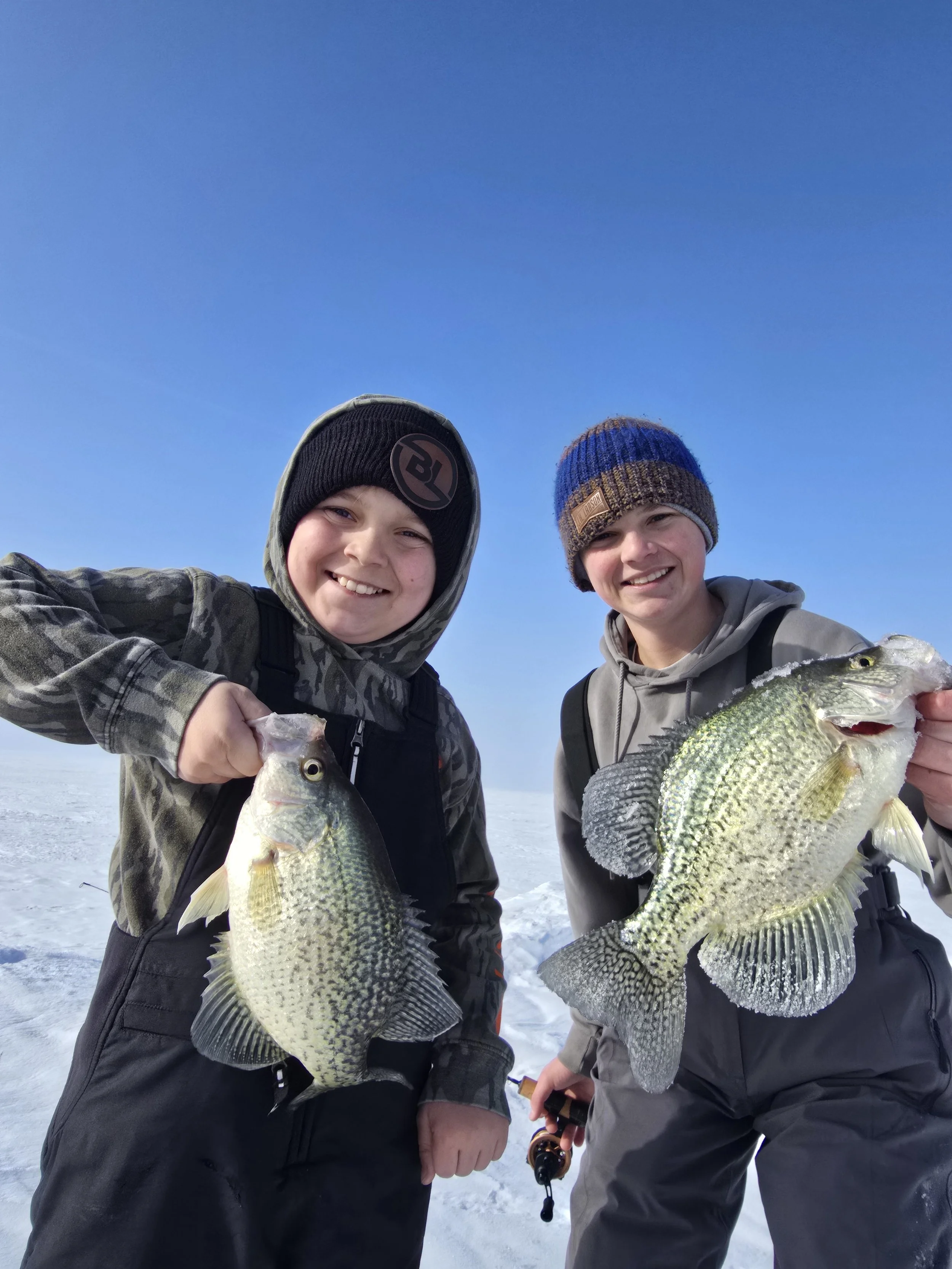 Two boys in winter clothing standing on snow holding large fish they caught, with a clear blue sky in the background.