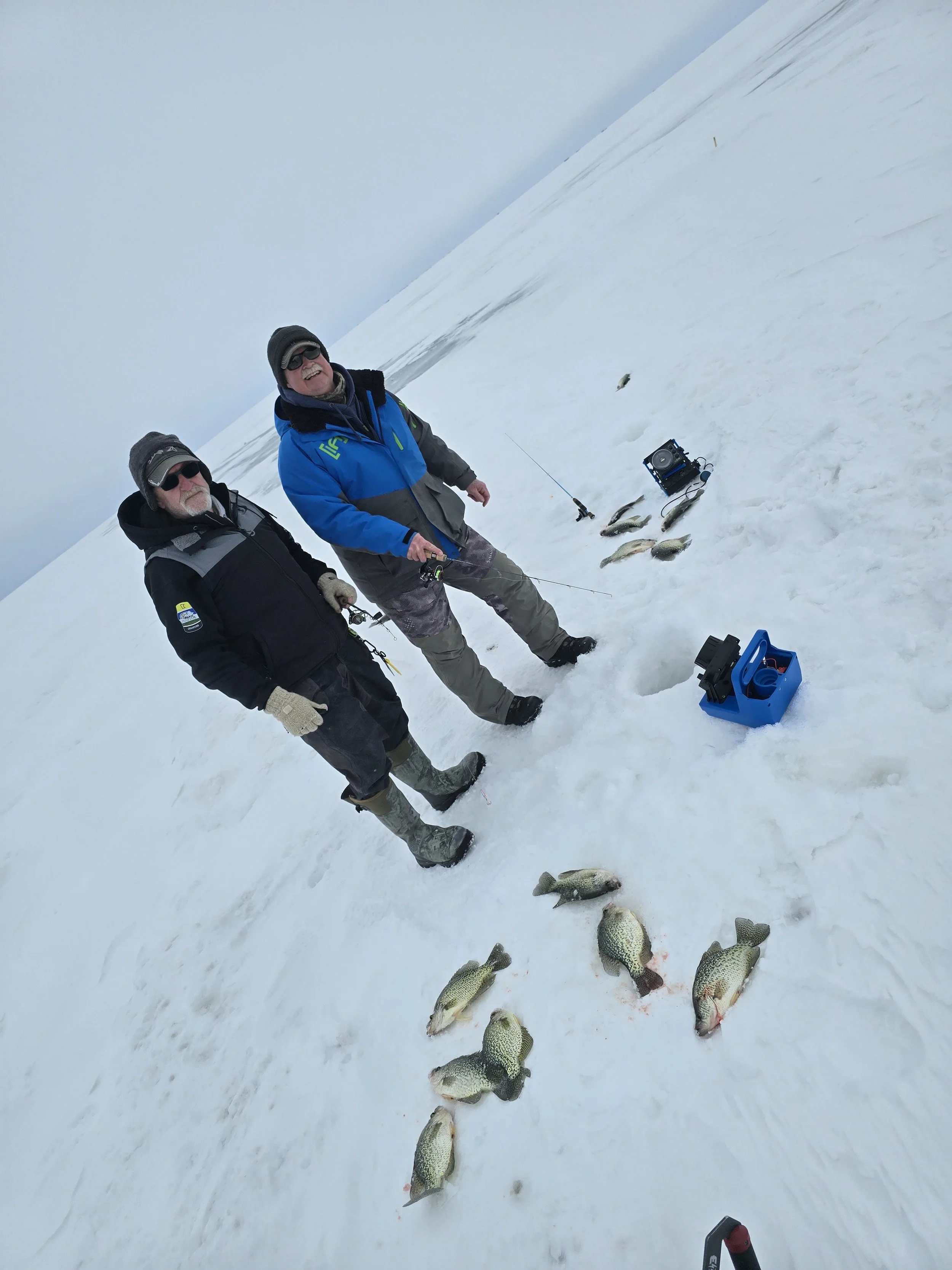 Two men ice fishing on a snowy landscape with several fish laid out on the snow and fishing equipment nearby.