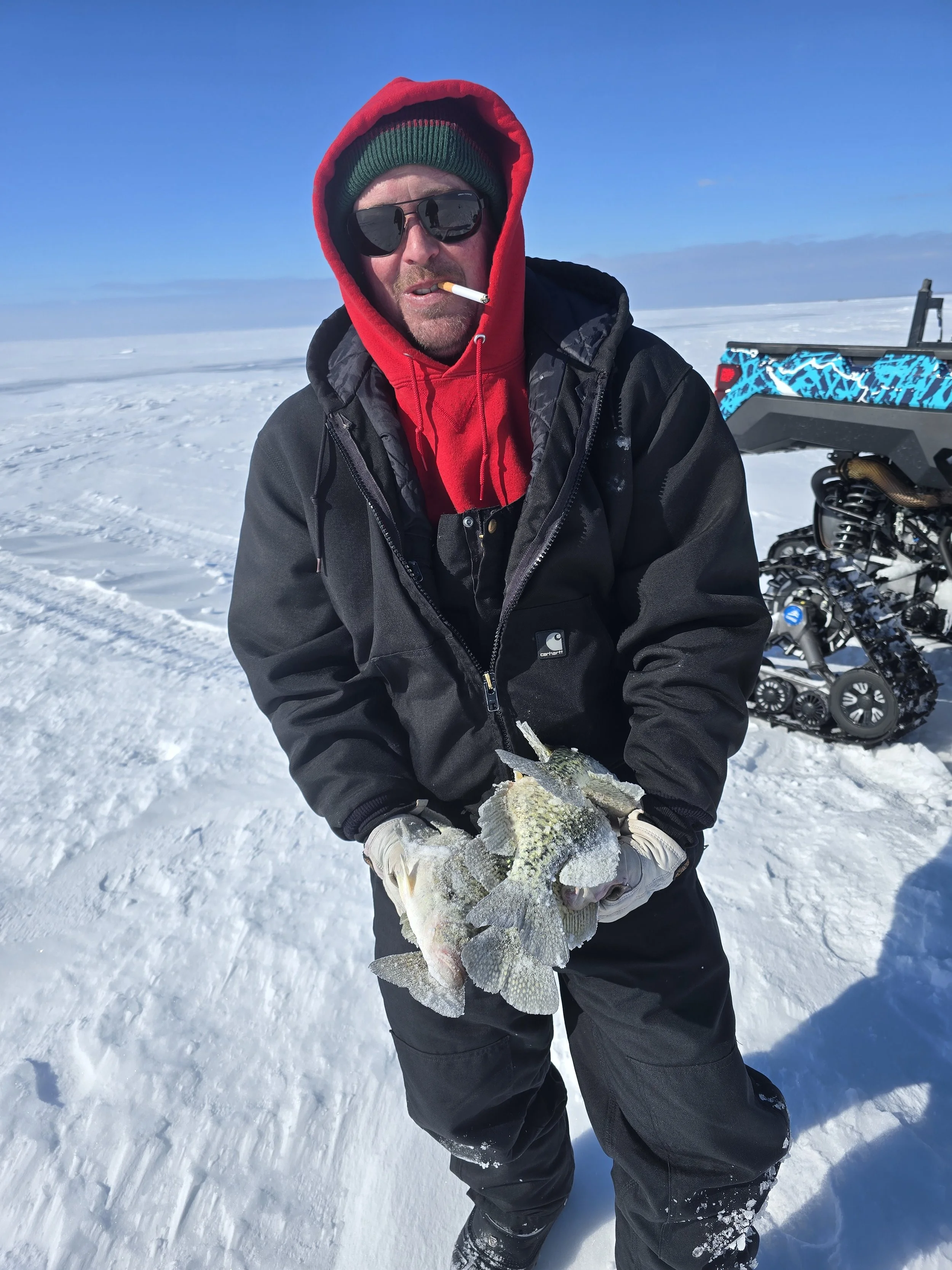 A man dressed in winter gear holding a large fish he caught, standing on snow with a snowmobile and a snowy landscape in the background.