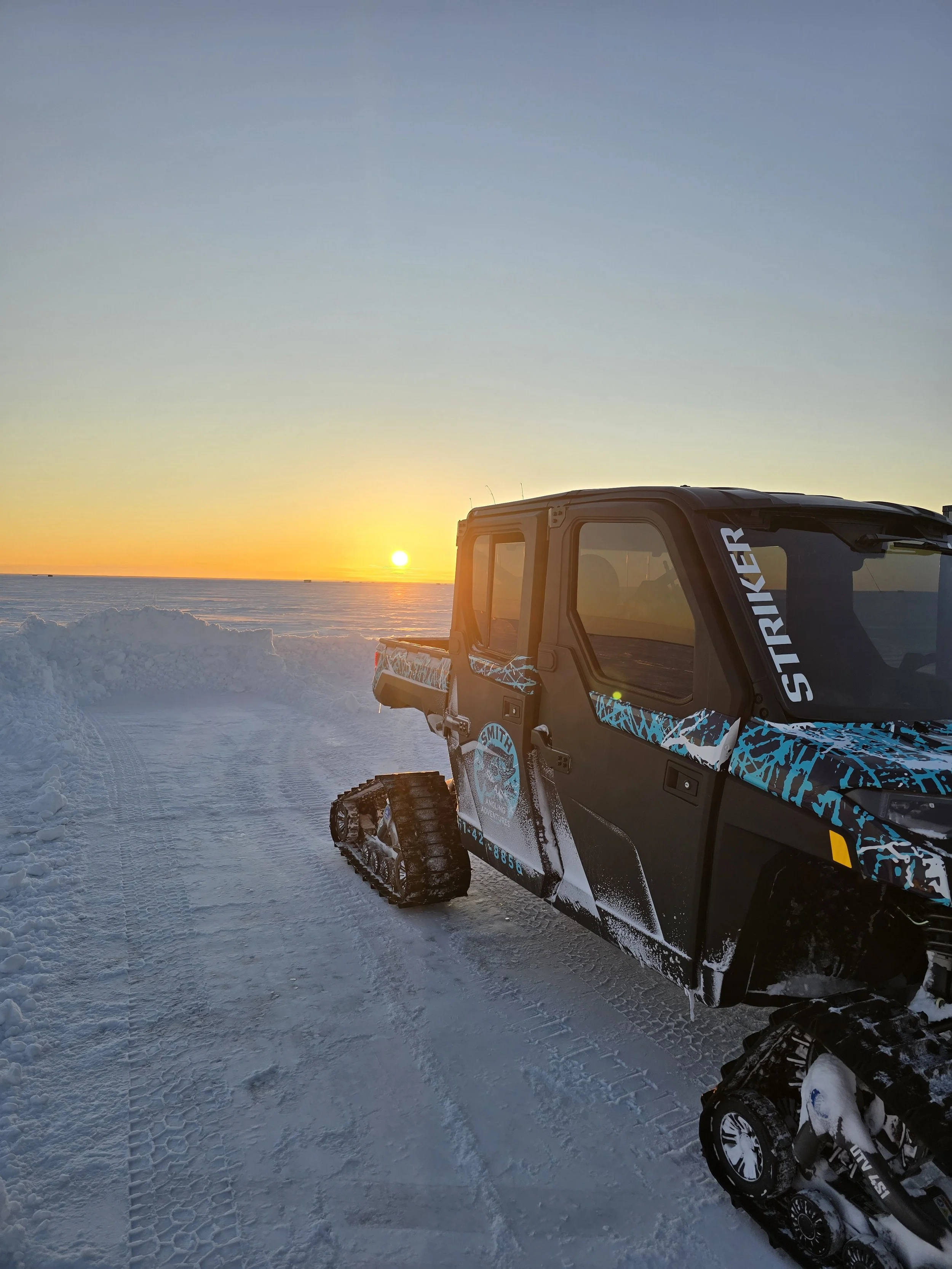 A snow-covered landscape at sunset with part of a tracked vehicle, likely an ATV or UTV, visible in the foreground. The sky shows a gradient from pale blue to orange near the horizon.