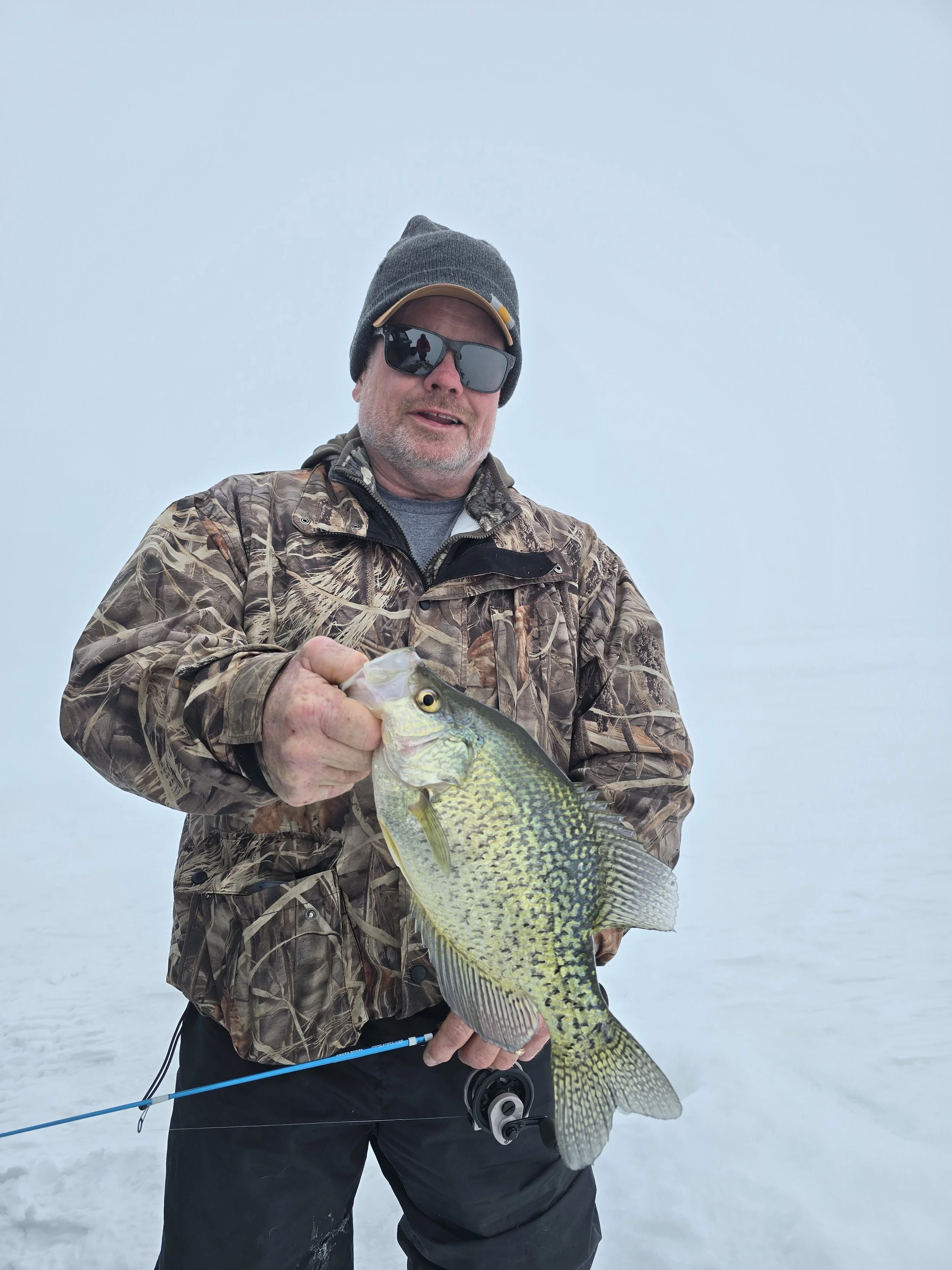 A man wearing a gray hat, sunglasses, camouflage jacket, and black pants, standing on a snowy surface, holding a large fish with yellow and green scales.
