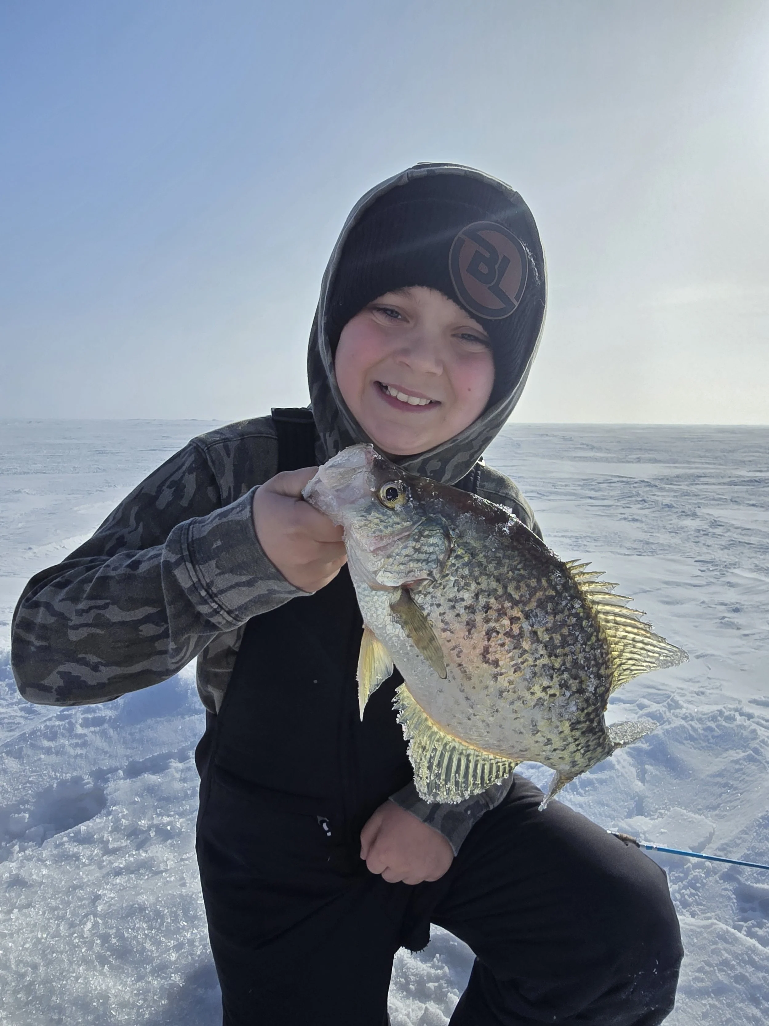 A young boy dressed in winter clothing holding a large fish on a snowy landscape with a clear sky.