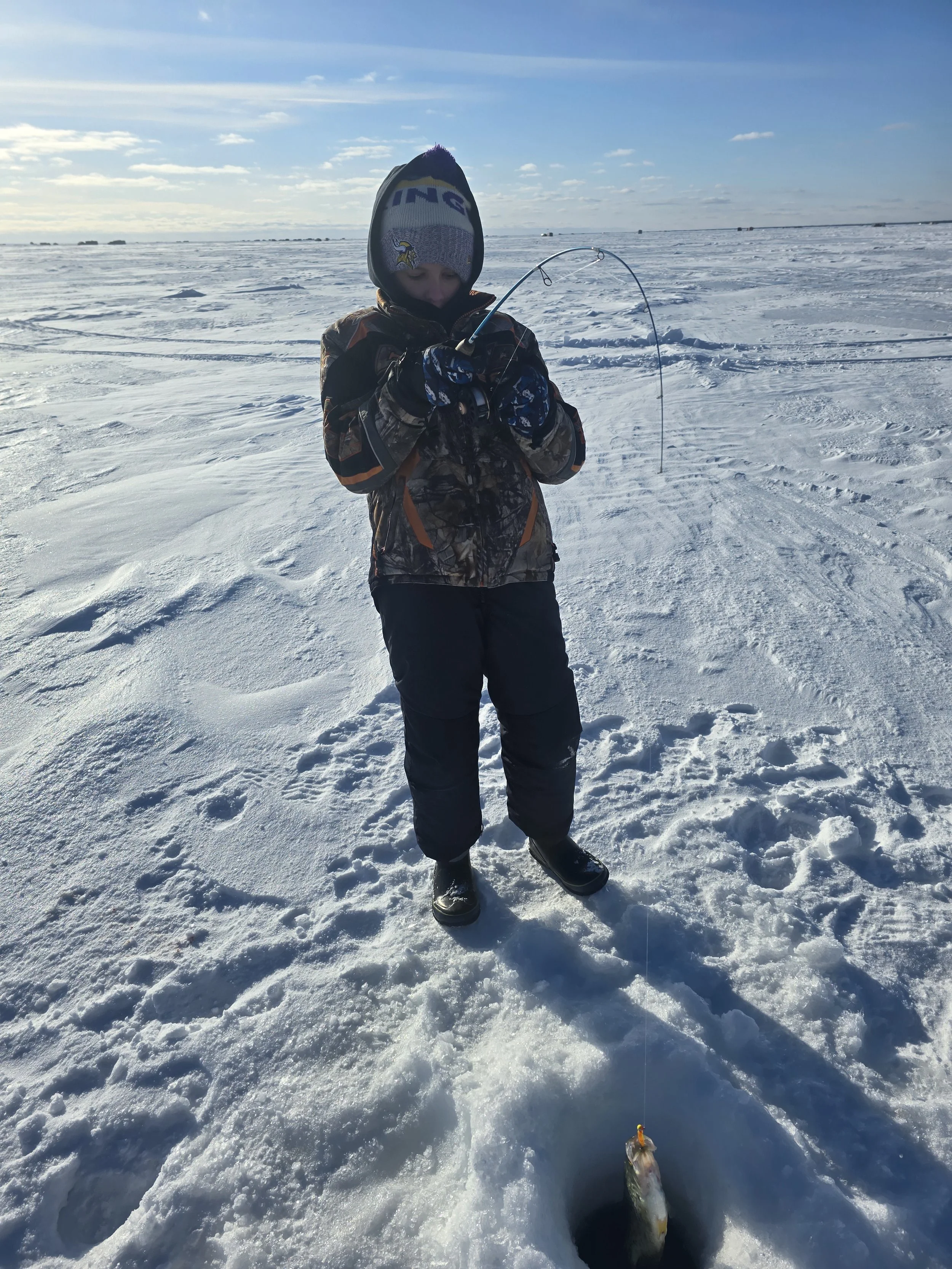 A young child wearing winter clothing, including a Minnesota Vikings beanie, standing on snow, holding a fishing rod with a fish caught on the line at a frozen lake.
