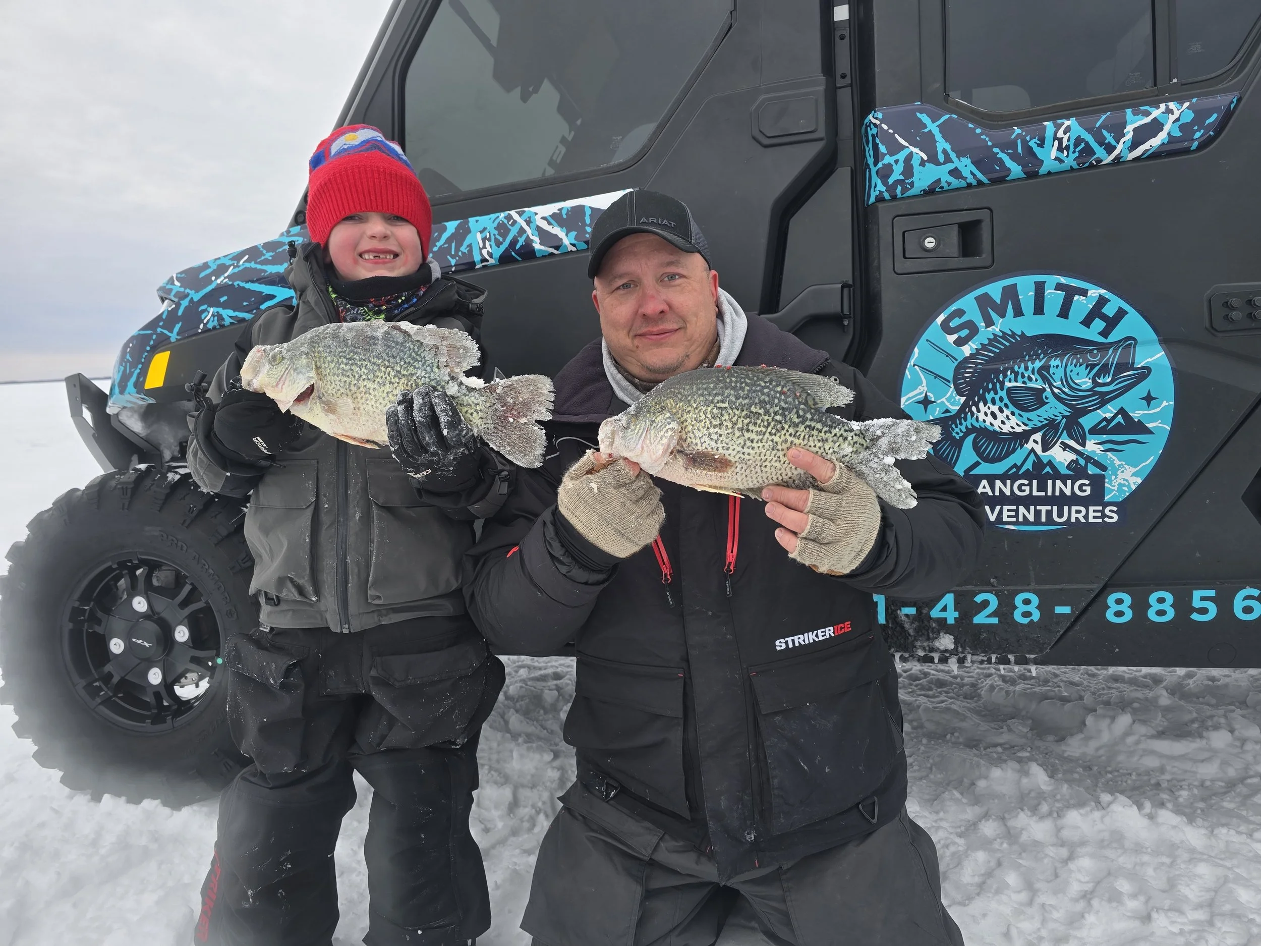 Two people standing in the snow holding large fish in front of a black vehicle with blue and white graphic decals. One person is a child wearing a red hat and black winter gear, and the other is an adult wearing a black cap, gray hoodie, and black wi