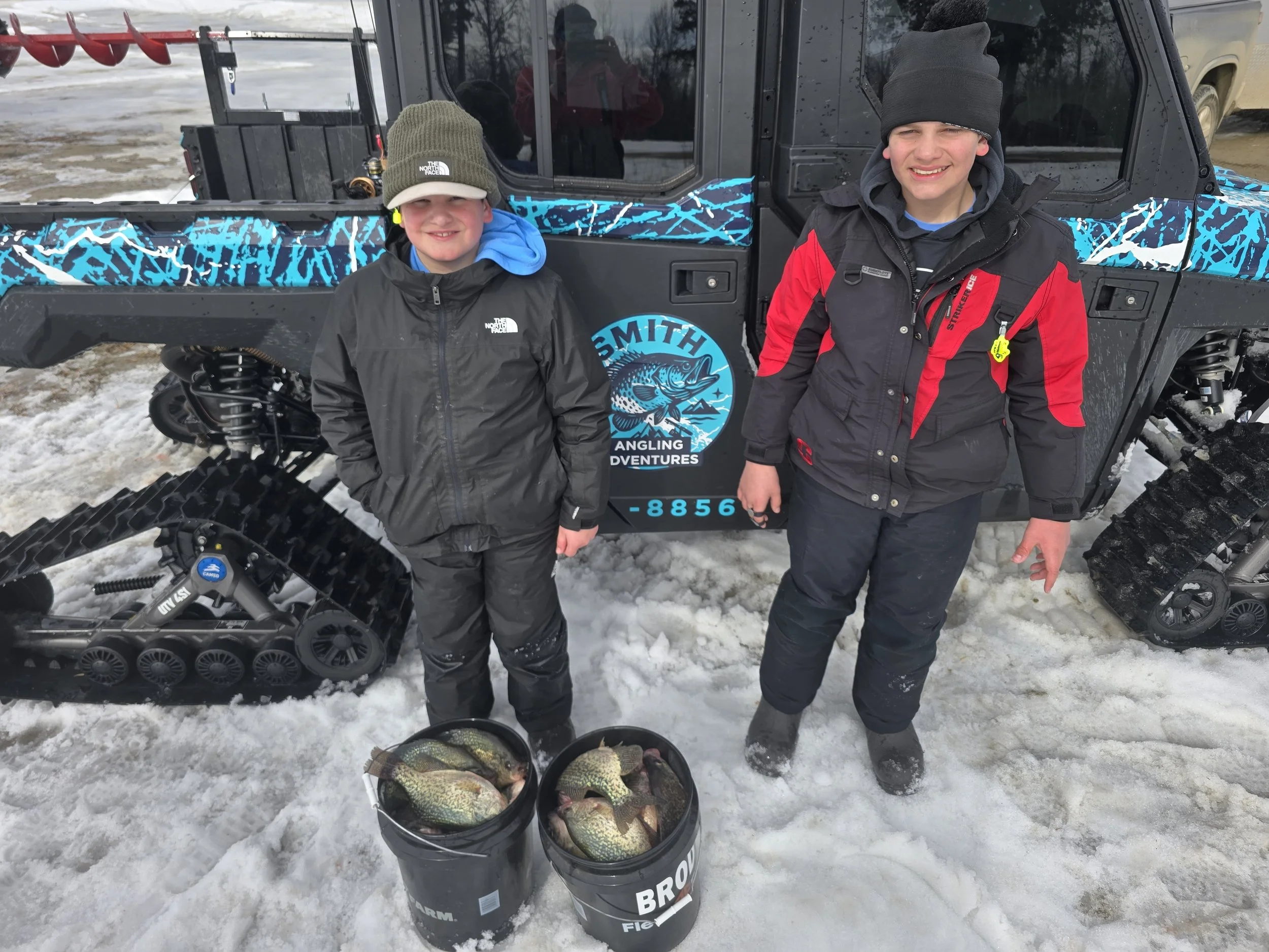 Two boys in winter clothing standing on snow with fishing equipment and holding buckets of fish in front of an off-road vehicle, outdoors on a snowy day.