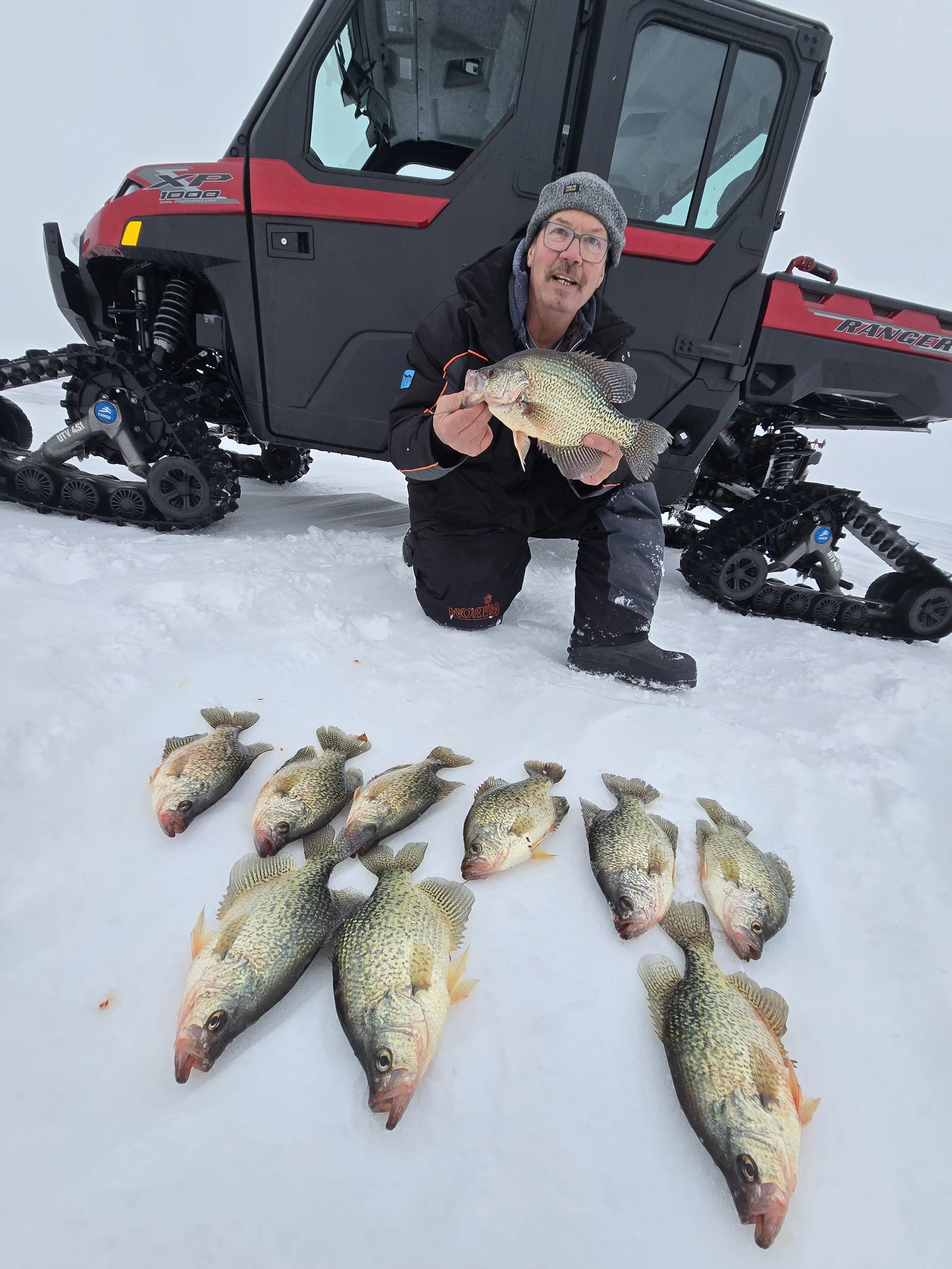 A man wearing winter clothing and glasses kneeling in the snow, holding a large fish. Several other fish are laid out on the snow in front of him. A snow-covered landscape with a red and black snowcat vehicle in the background.