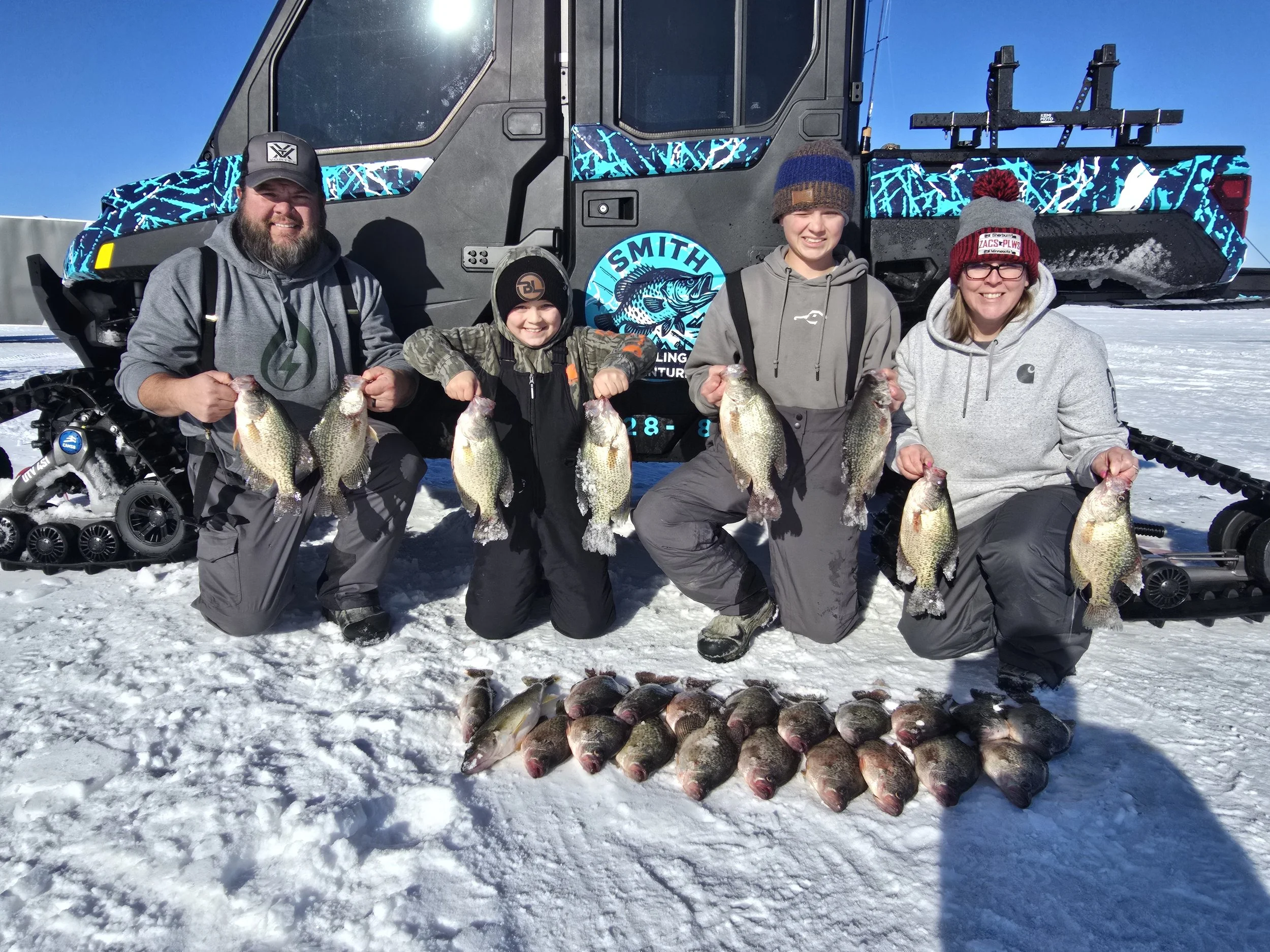 A group of four people standing on snow, smiling and holding large fish they caught, with a snowmobile and a decorated vehicle behind them.