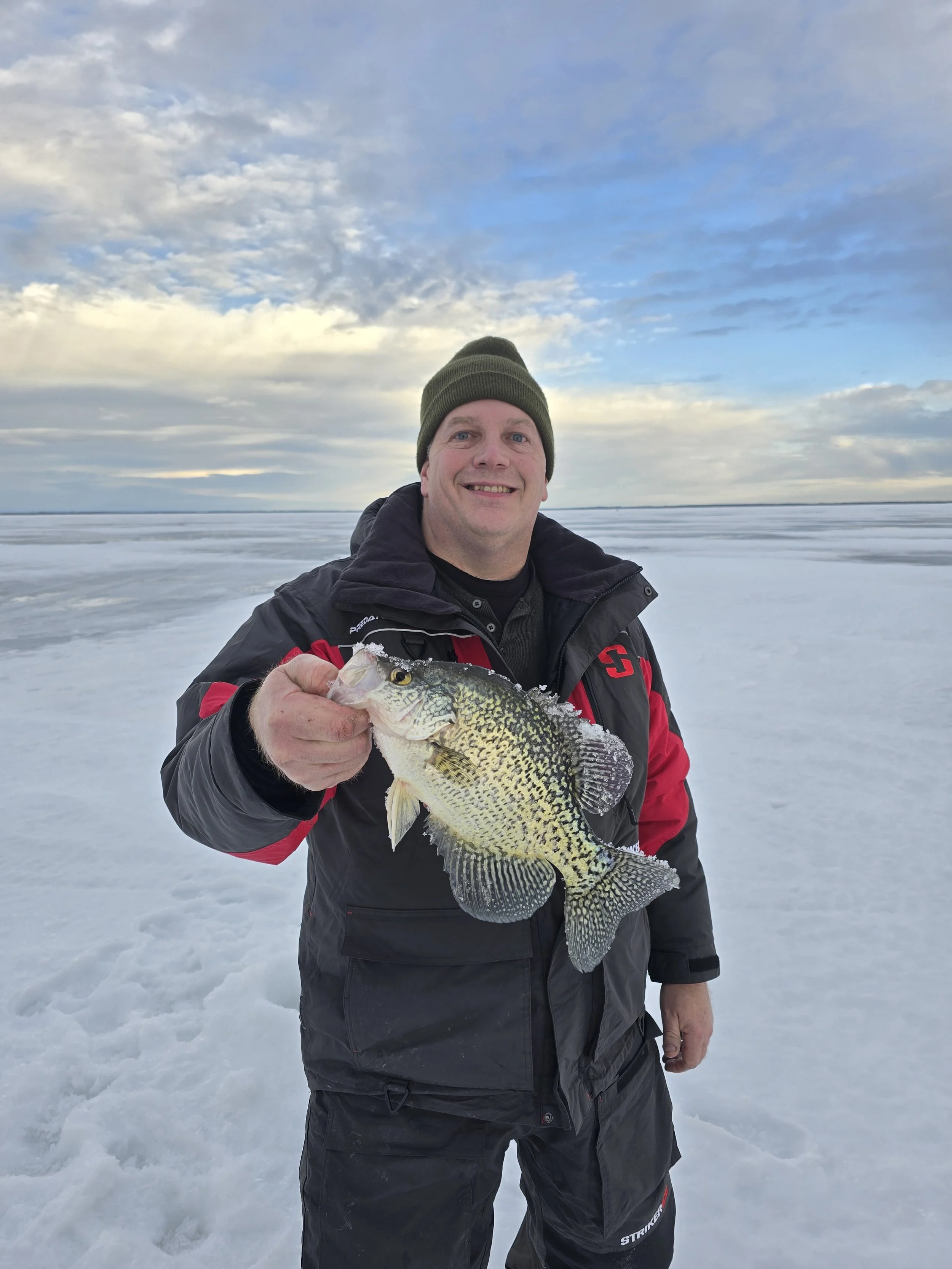 A man in winter clothing holding a fish on a snowy frozen lake with a partly cloudy sky in the background.