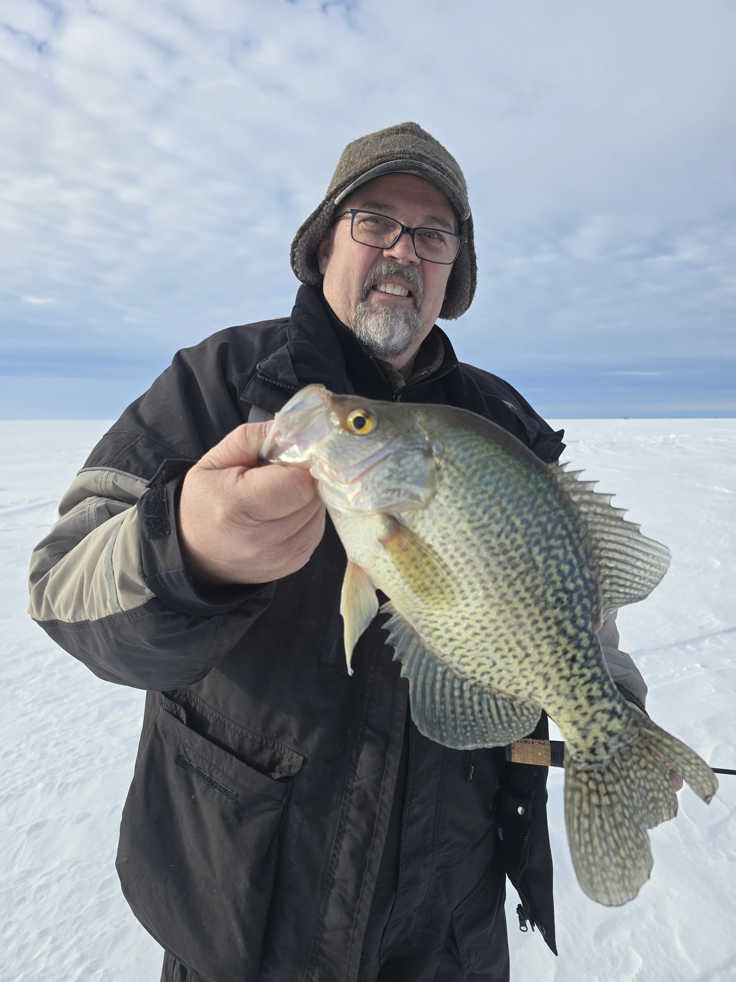 A man outdoors in winter holding a fish in front of him, with snow and a cloudy sky in the background.
