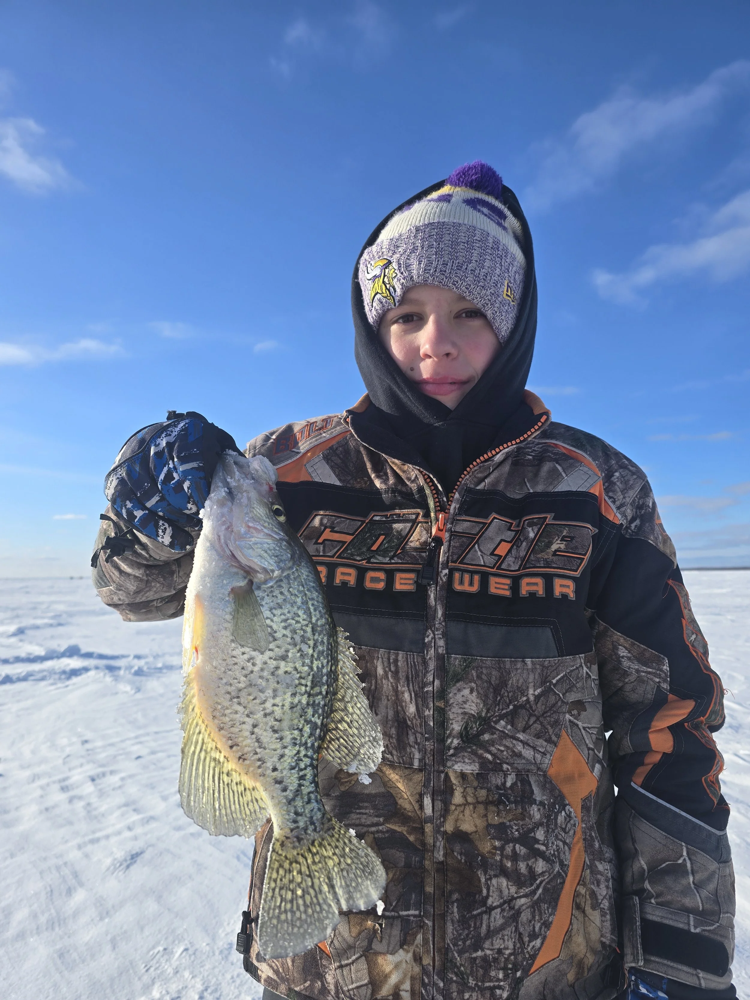 A young boy dressed in winter camouflage gear holding a fish in a snowy, outdoor landscape with a blue sky and few clouds.