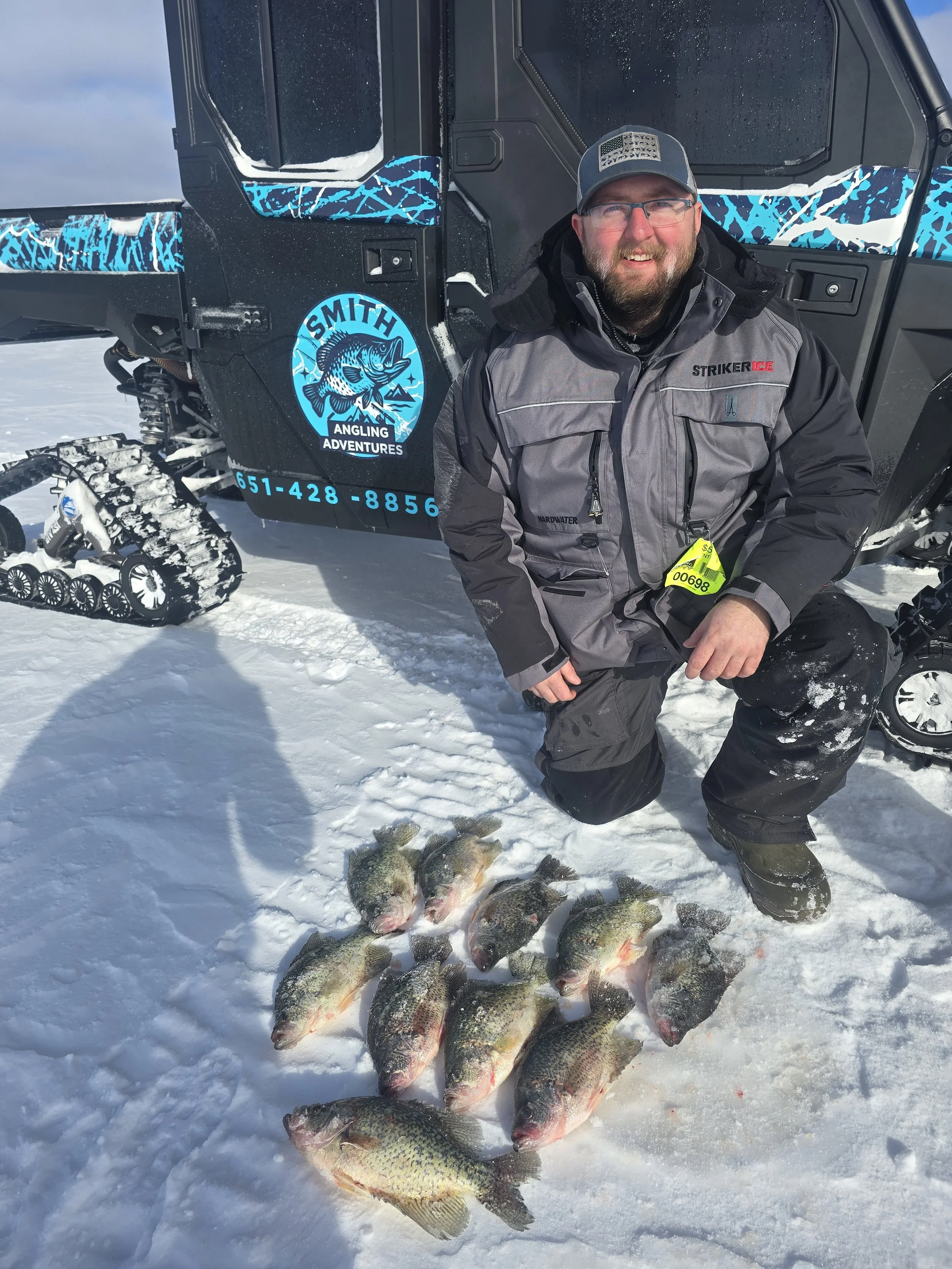 A man dressed in winter gear kneeling on snow in front of an ice fishing vehicle, with several large fish laid out on the snow, and a tracked snowmobile in the background.