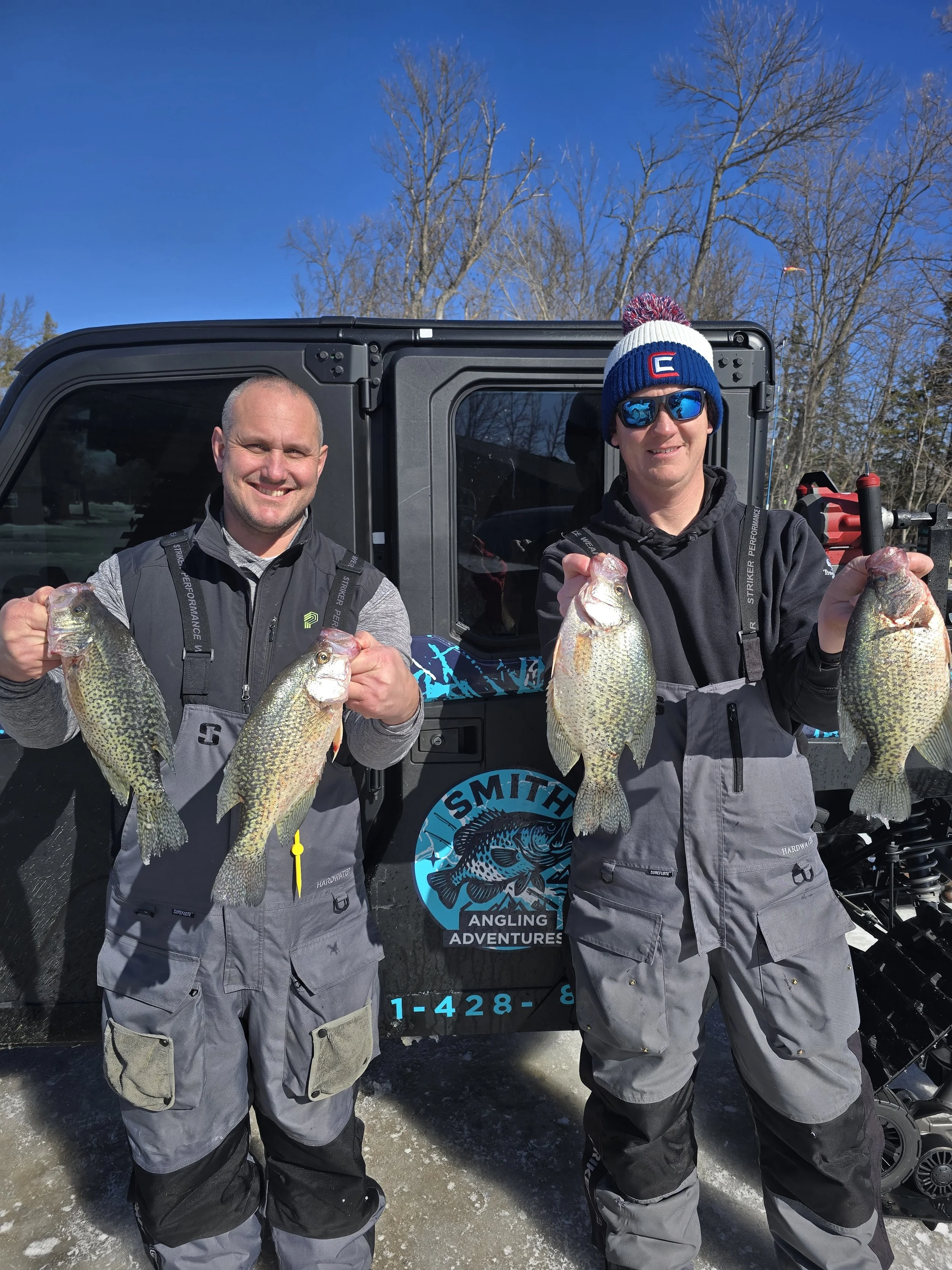 Two men smiling and holding large fish in front of a vehicle with an angling adventure logo. They are dressed in fishing gear, outdoors on a sunny day with bare trees in the background.