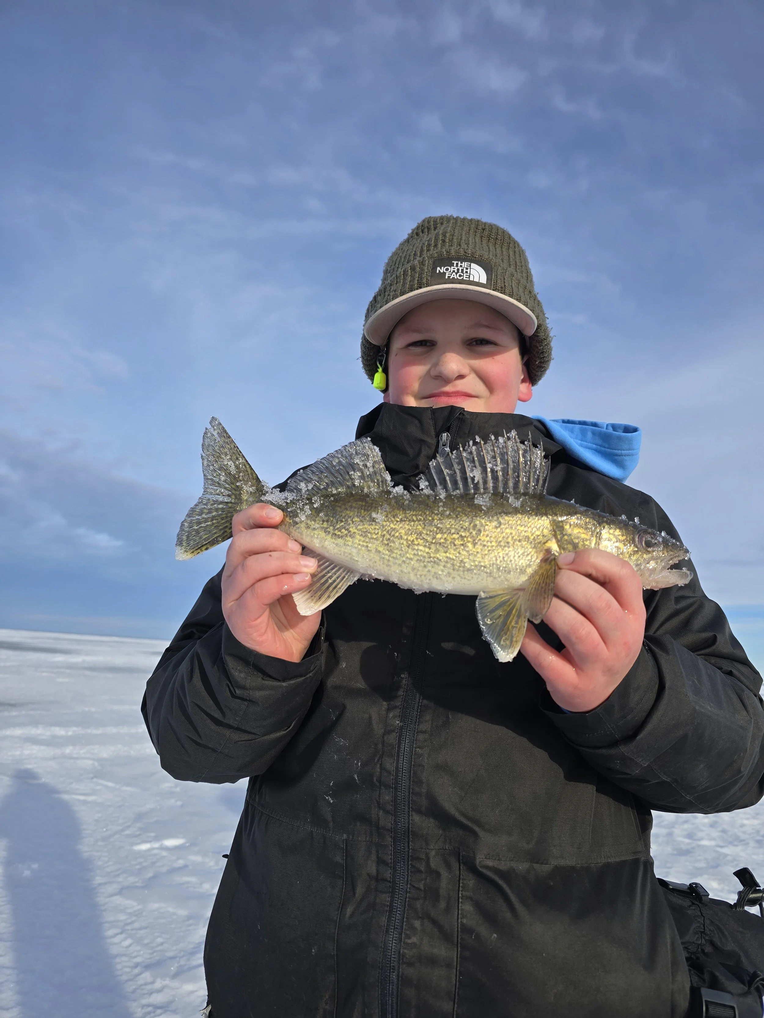 A young boy wearing a gray beanie and a black jacket, standing on snow, holding a fish with yellow and gray coloring, with a blue sky in the background.