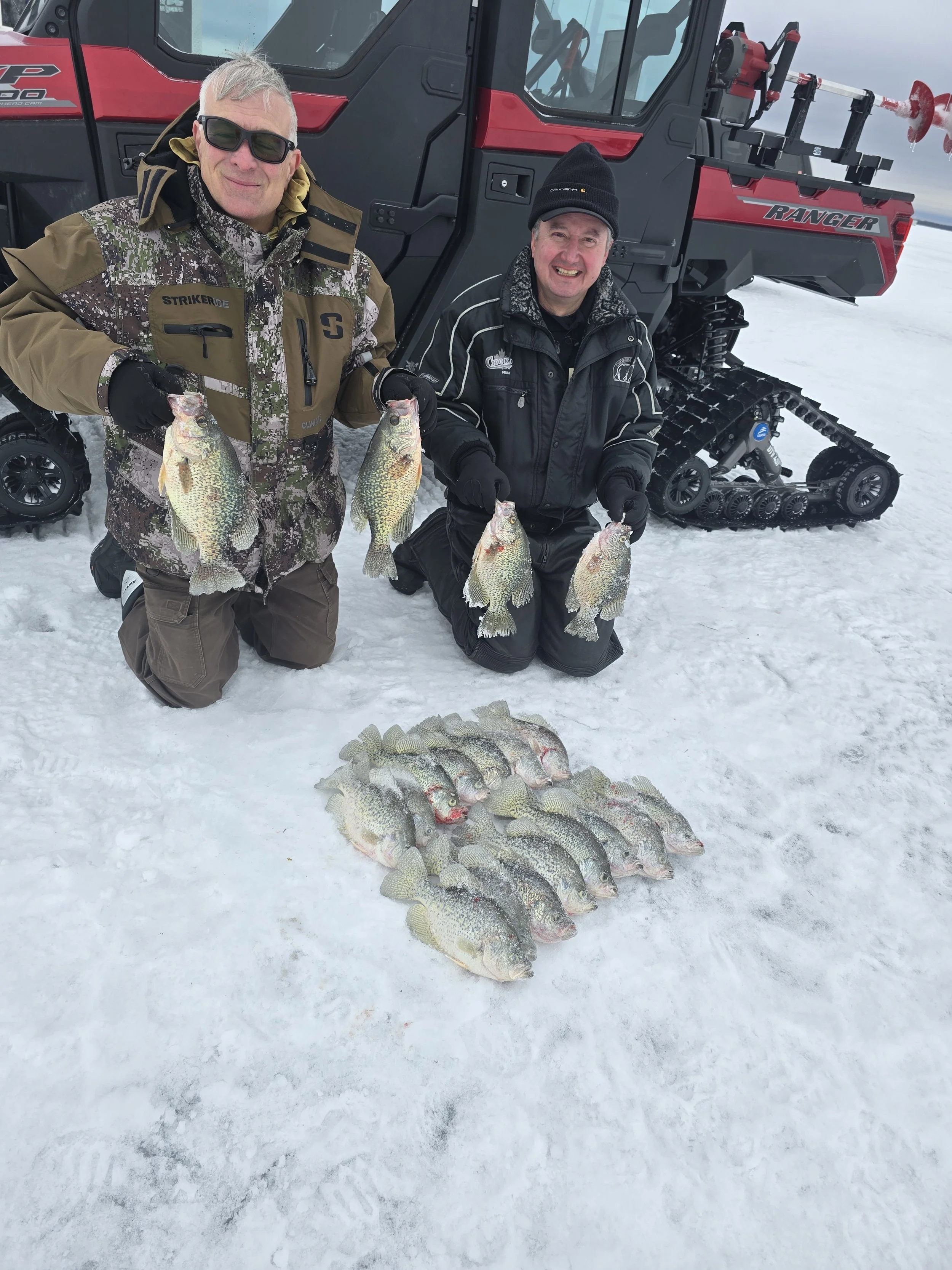 Two men kneeling on snow-covered ground holding freshly caught fish, with a tracked vehicle for snow and ice in the background.