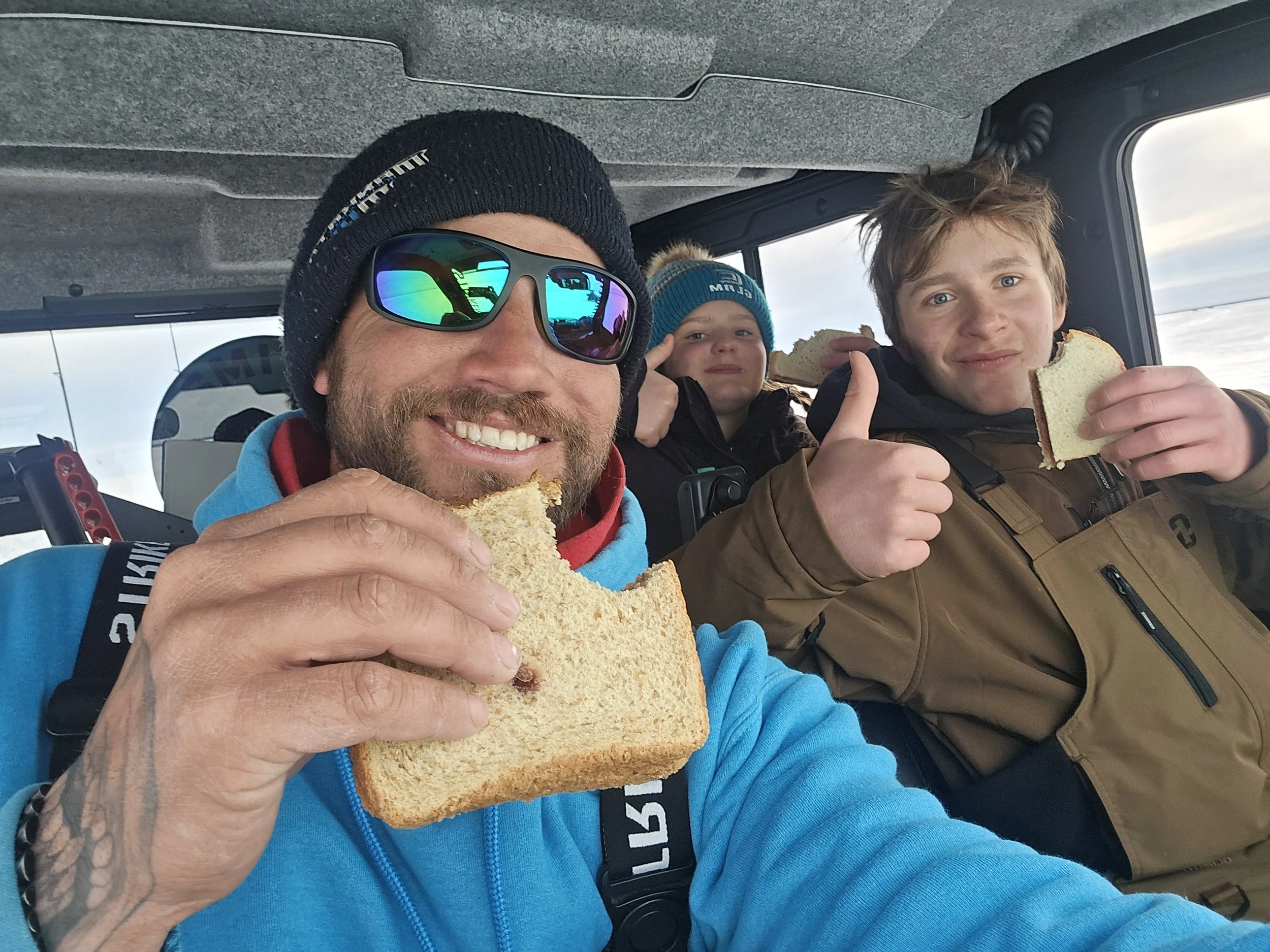 A man with sunglasses and a knit hat smiling while holding a sandwich, sitting with two children, one of whom is also eating a sandwich, inside a vehicle with cloudy skies visible outside.