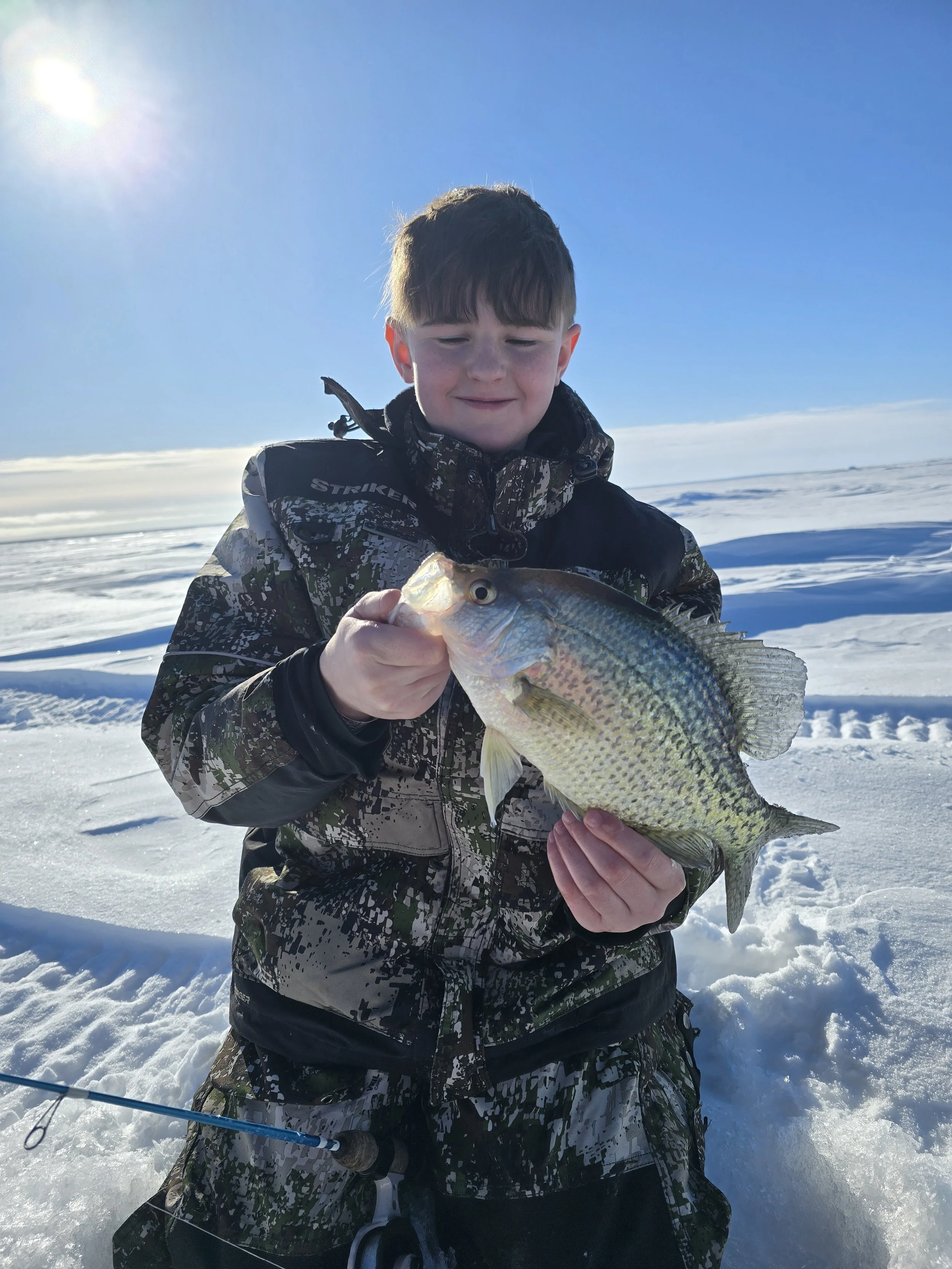 A young boy wearing camouflage winter clothing kneeling on snow-covered ground holding a fish with both hands.