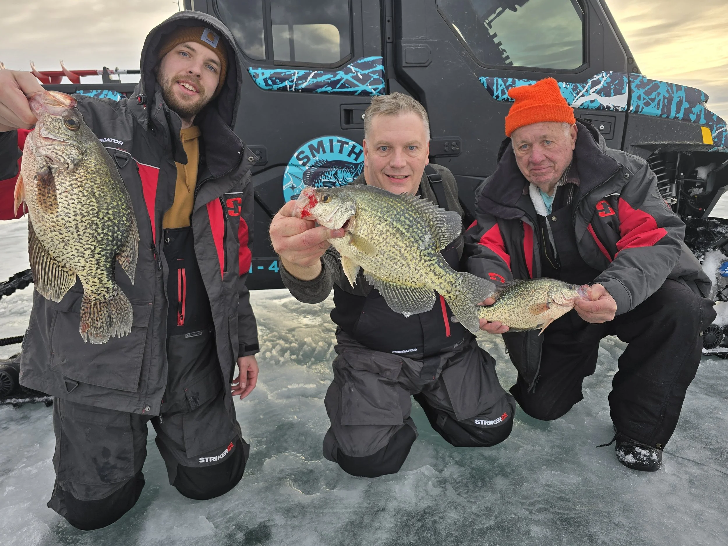 Three men in winter gear on an icy lake holding fish they caught, with a snowmobile behind them and a cloudy sky above.
