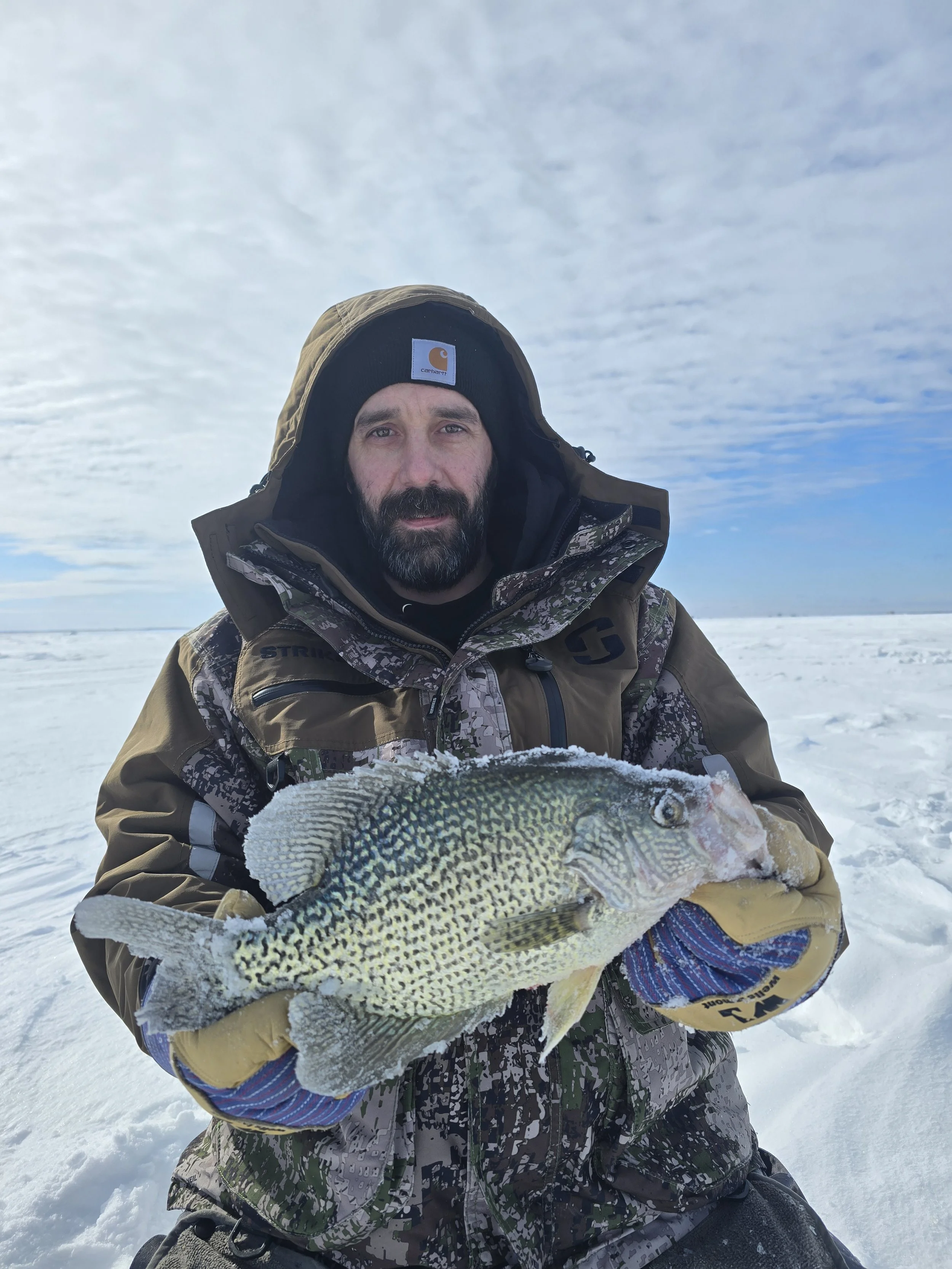 Man in camouflage jacket and black Carhartt beanie holding a large fish on snow-covered ground, with cloudy sky in background.