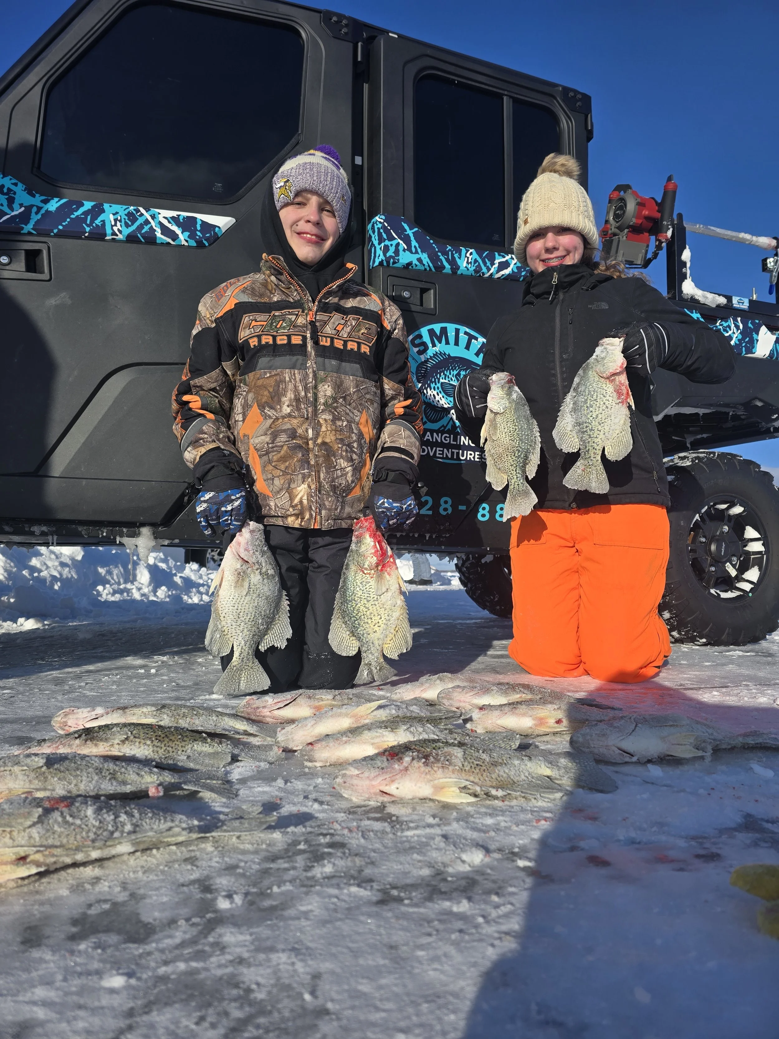 Two children standing on snow holding fish in winter clothing in front of a black off-road vehicle with a blue and black design and logo, with other fish laid out on the snow in front of them.