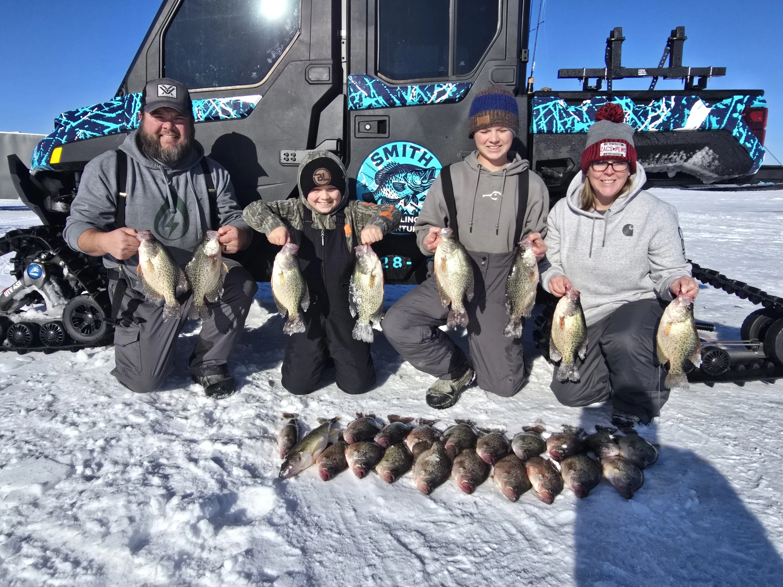 A group of five people, including two children and three adults, standing on snow in front of a snowmobile and a large snowmobile trailer. They are holding up large fish they caught during a winter fishing trip, with a pile of smaller fish on the sno
